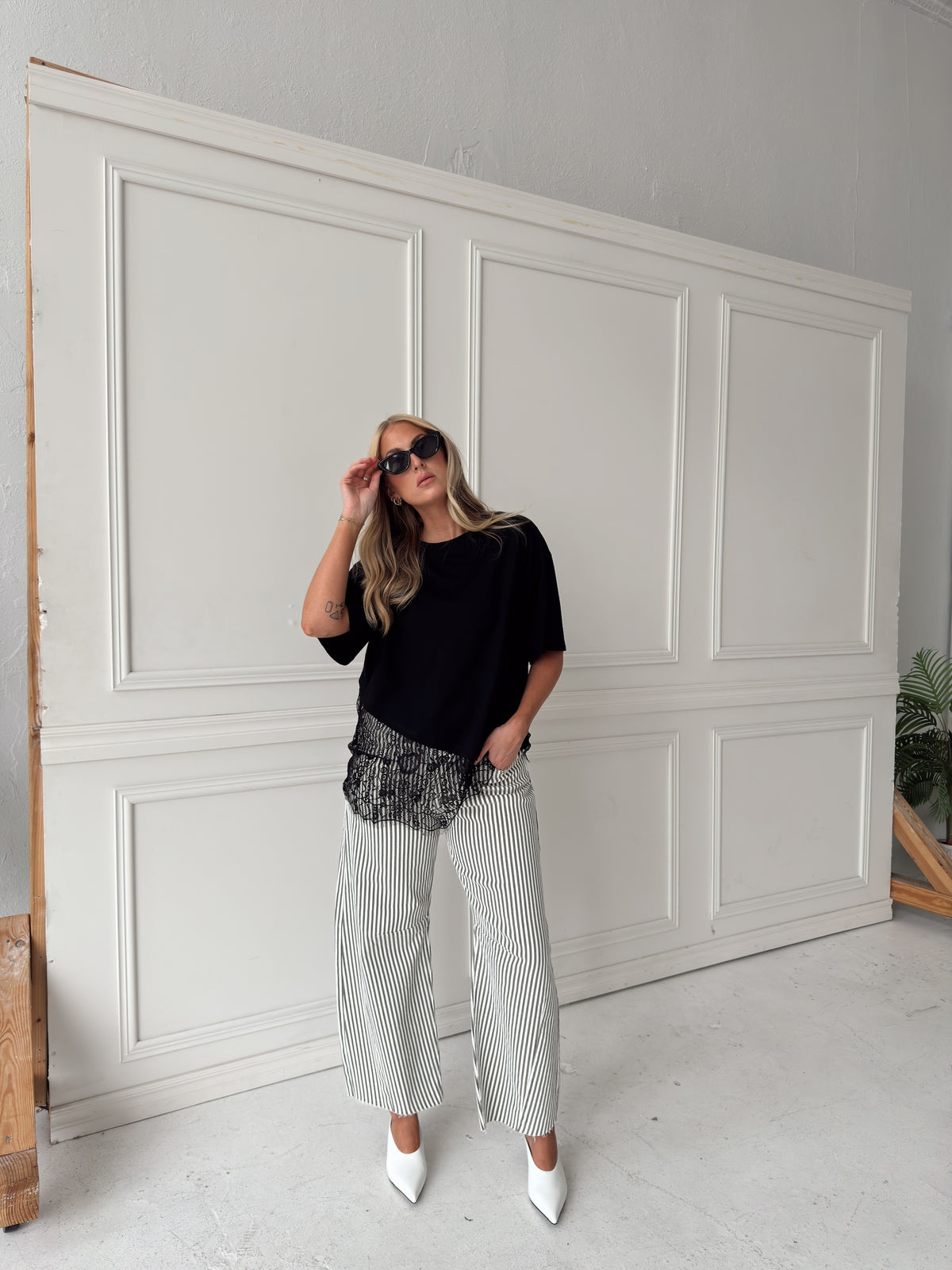 A woman modeling a pair of wide-leg barrel jeans with thin olive and white vertical stripes, paired with a black t-shirt, black sunglasses, and white pointed-toe heels, standing in front of a white paneled wall.