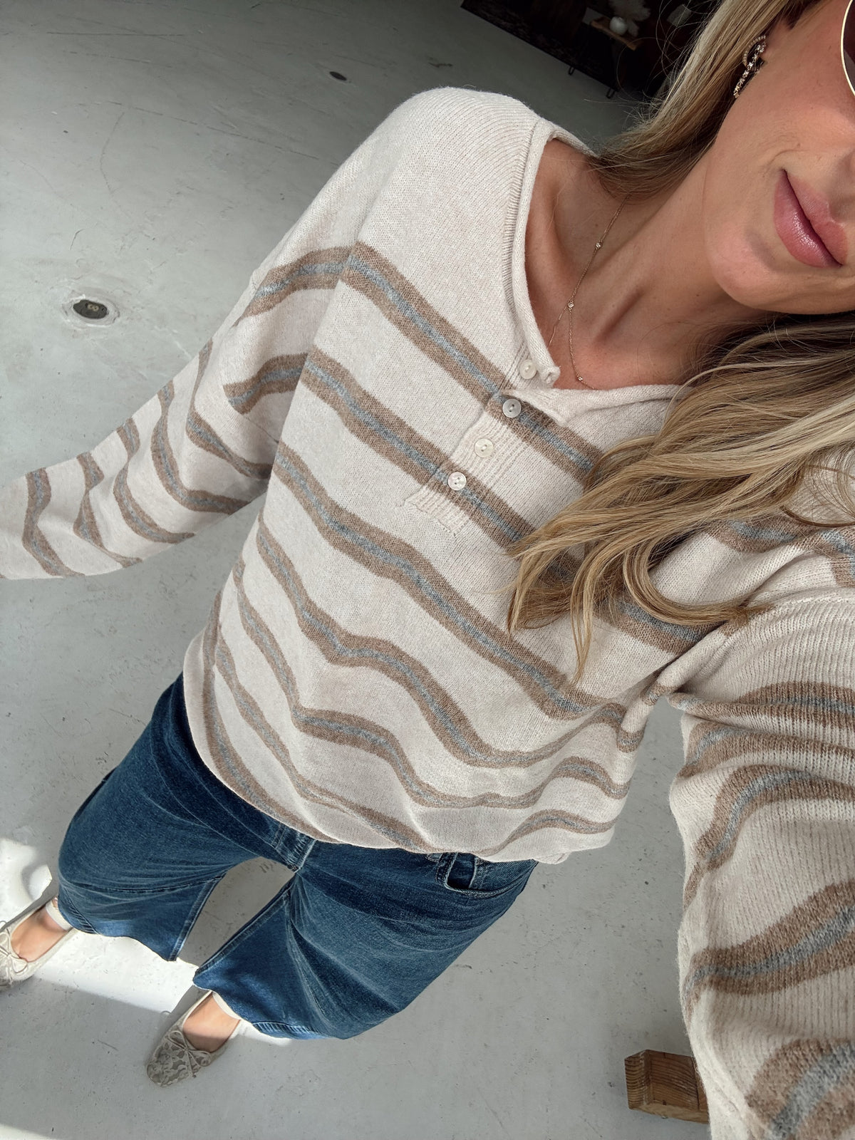 A woman wearing a beige, cream, and light blue striped knit henley sweater, blue jeans, and light colored slip-on shoes, captured in a casual indoor selfie.