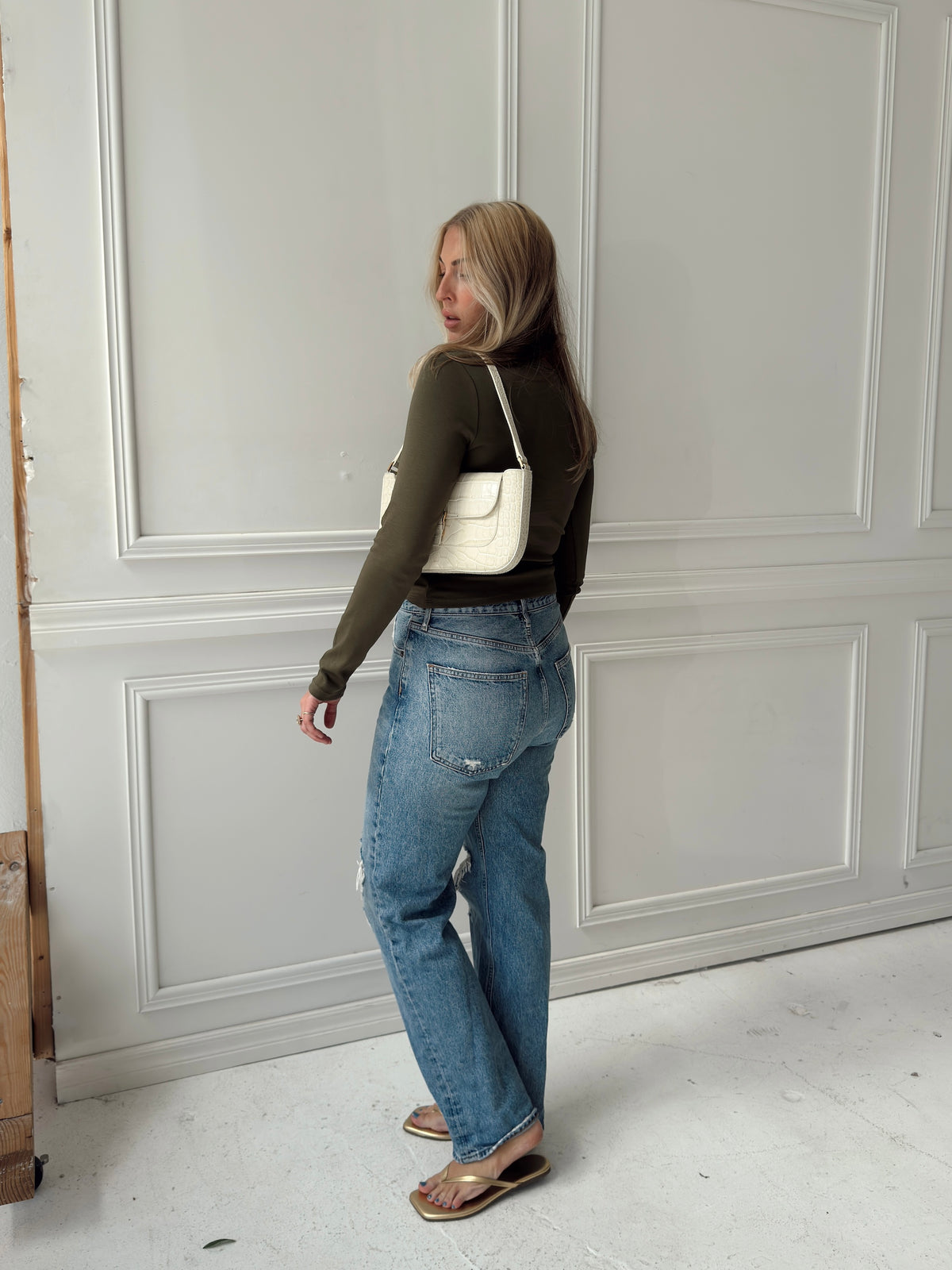 A woman posing in a studio wearing an olive green long-sleeve top and light-wash blue jeans with distressed details.