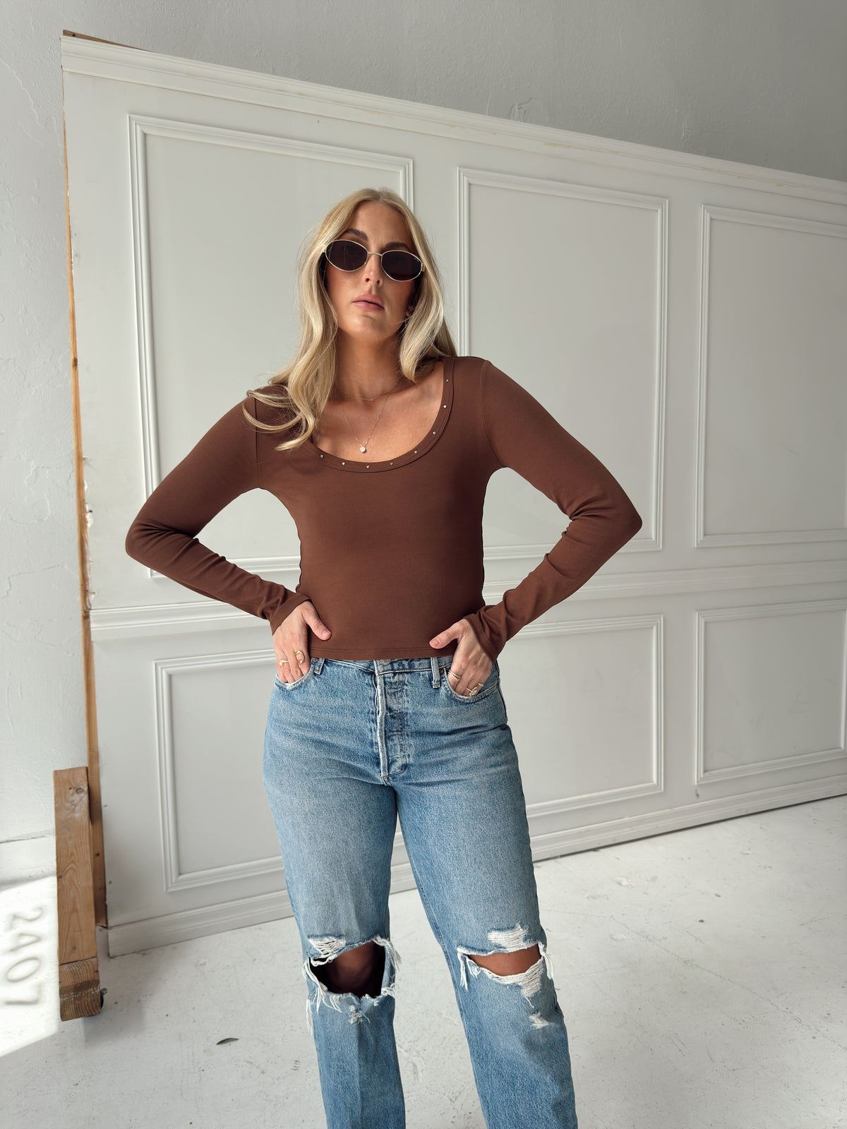 A woman modeling a brown long-sleeve scoop-neck top with subtle rhinestone trim along the neckline, paired with distressed blue jeans, standing in front of a white paneled wall.