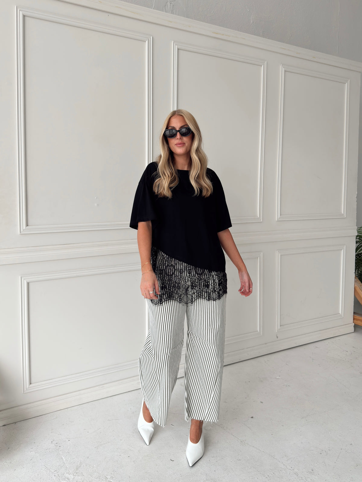 A woman modeling a black asymmetrical top with a sheer black lace hem, paired with white and olive vertically striped pants, standing in front of a white paneled studio backdrop.