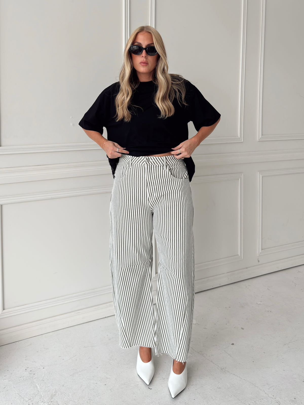 A woman modeling a pair of wide-leg barrel jeans with thin olive and white vertical stripes, paired with a black t-shirt, black sunglasses, and white pointed-toe heels, standing in front of a white paneled wall.