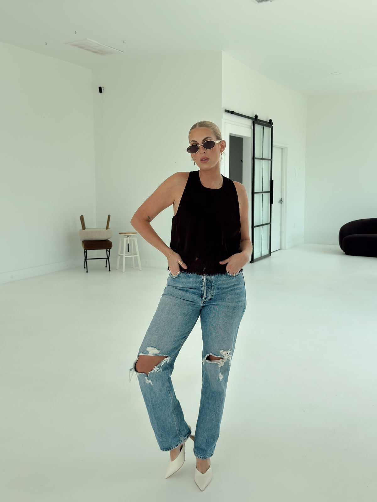 A woman posing in a minimalist white studio setting, wearing a brown sleeveless top, light-wash distressed blue jeans, and white pointed-toe heels.