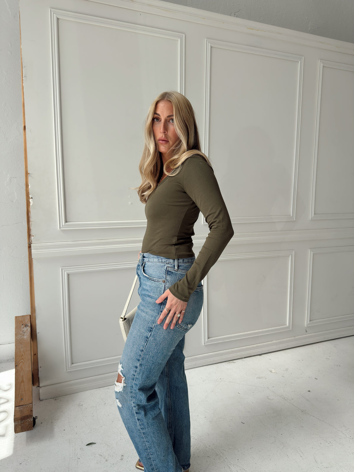 A woman posing in a studio wearing an olive green long-sleeve top and light-wash blue jeans with distressed details.