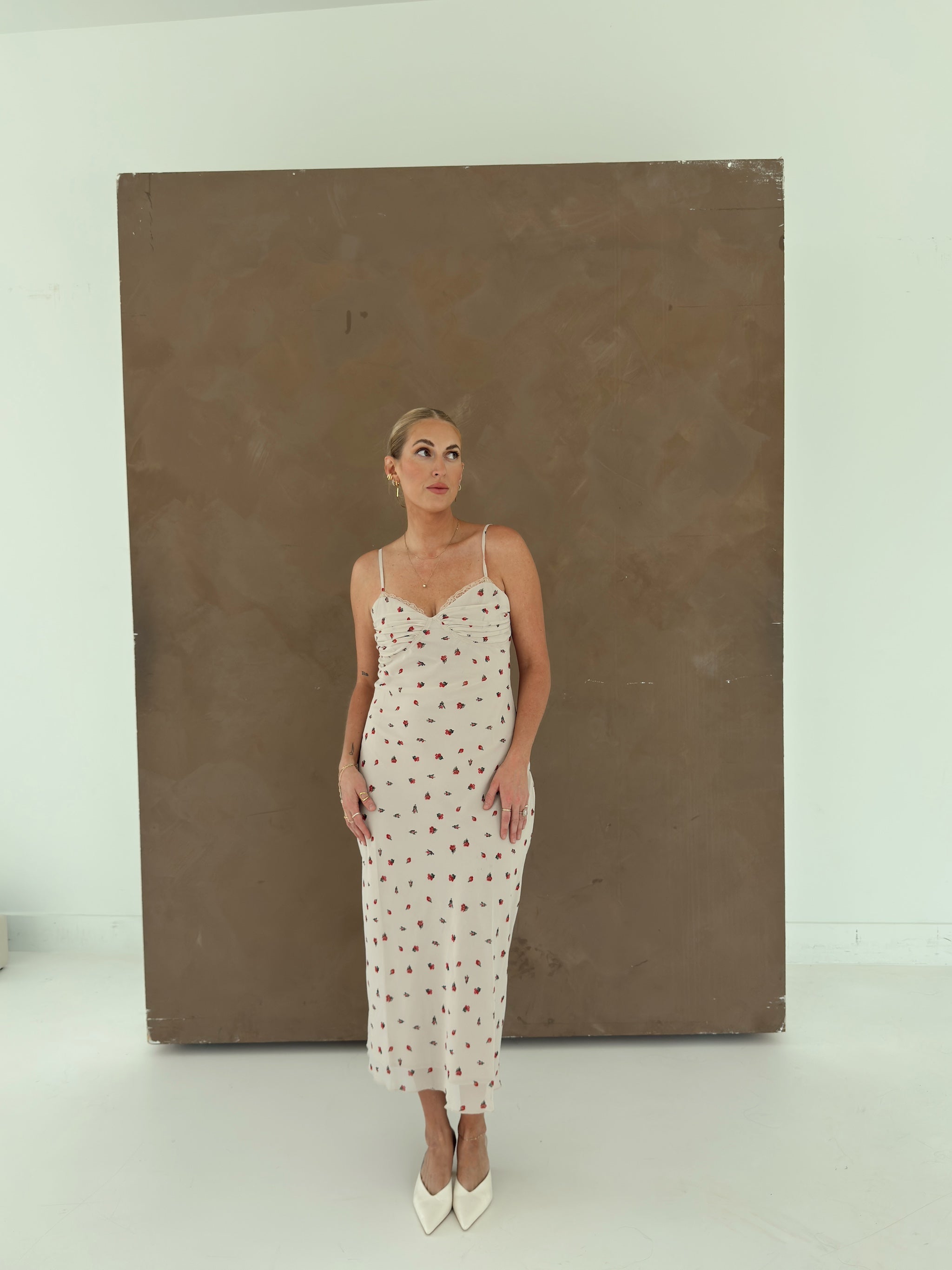 A model wearing a beige spaghetti-strap slip dress with small red ditsy floral, standing in front of a large brown canvas painting in a minimalist studio setting.