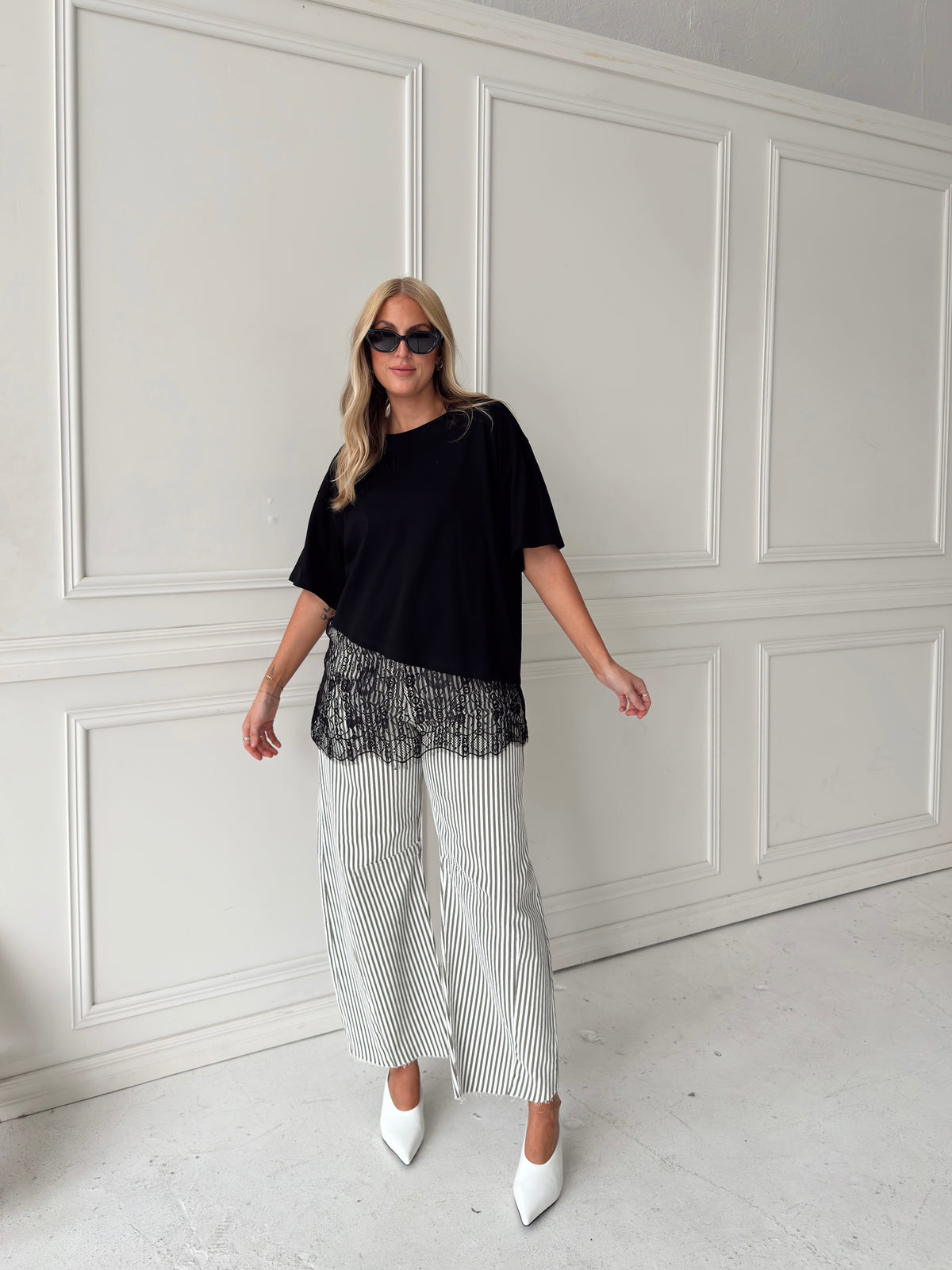 A woman modeling a black asymmetrical top with a sheer black lace hem, paired with white and olive vertically striped pants, standing in front of a white paneled studio backdrop.