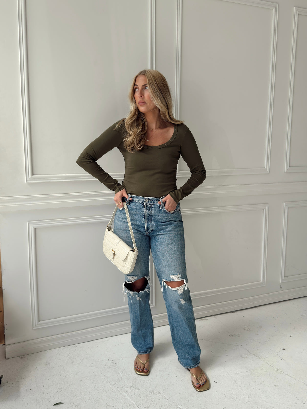 A woman stands in a room with white paneled walls wearing a fitted olive green long-sleeve top, distressed light-wash blue jeans with ripped knees, and holding a small white shoulder bag.