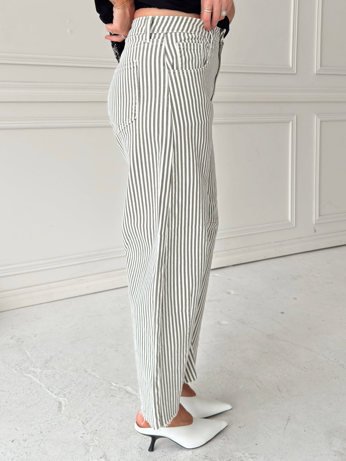 A person wearing high-waisted, wide-leg striped pants with vertical olive and white pinstripes, paired with white pointed-toe mule heels, standing in front of a white paneled wall.