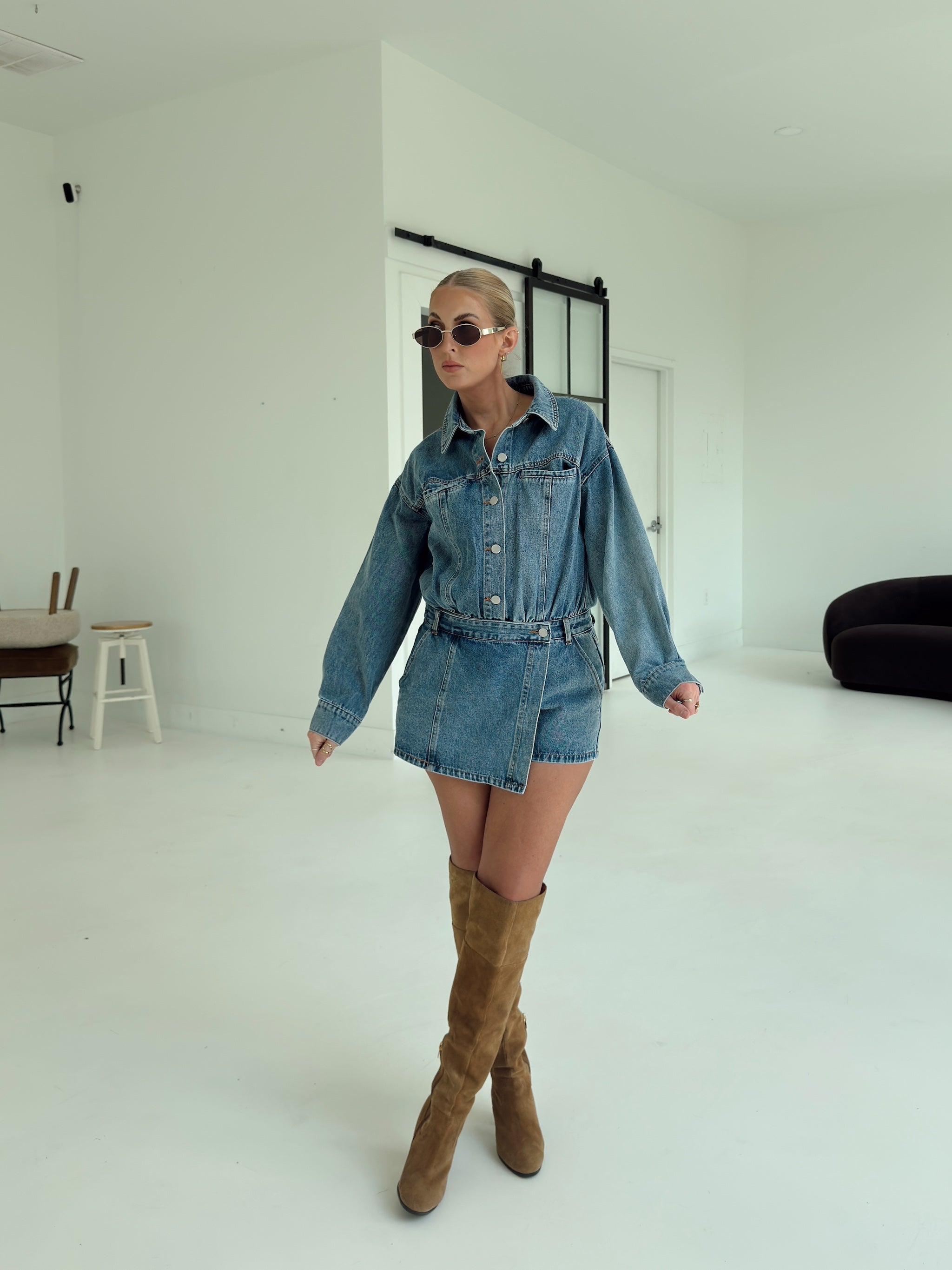 A woman modeling a blue denim romper, paired with brown suede over-the-knee boots, in a bright white studio setting.