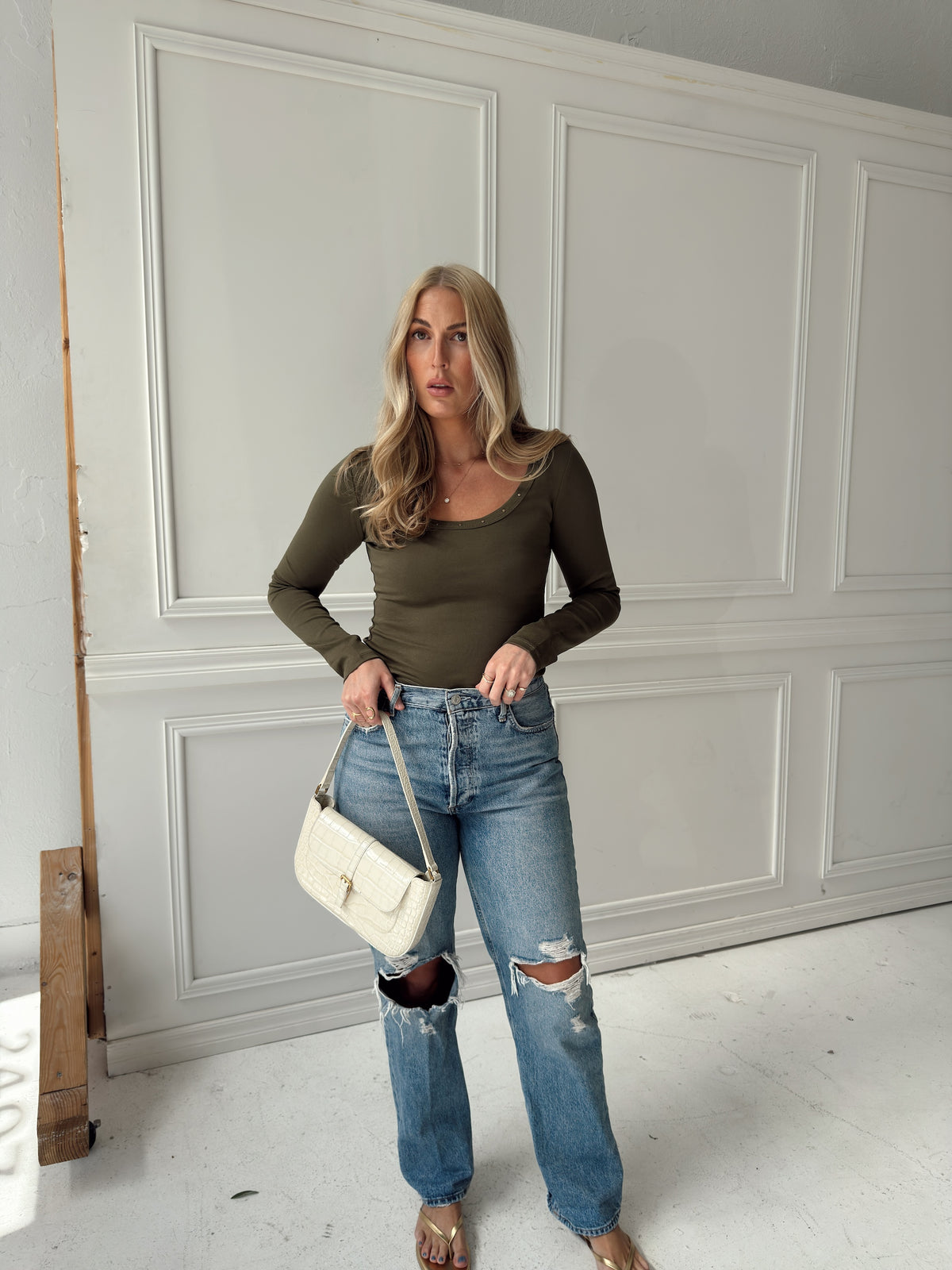 A woman stands in a room with white paneled walls wearing a fitted olive green long-sleeve top, distressed blue jeans, and holding a white shoulder bag.