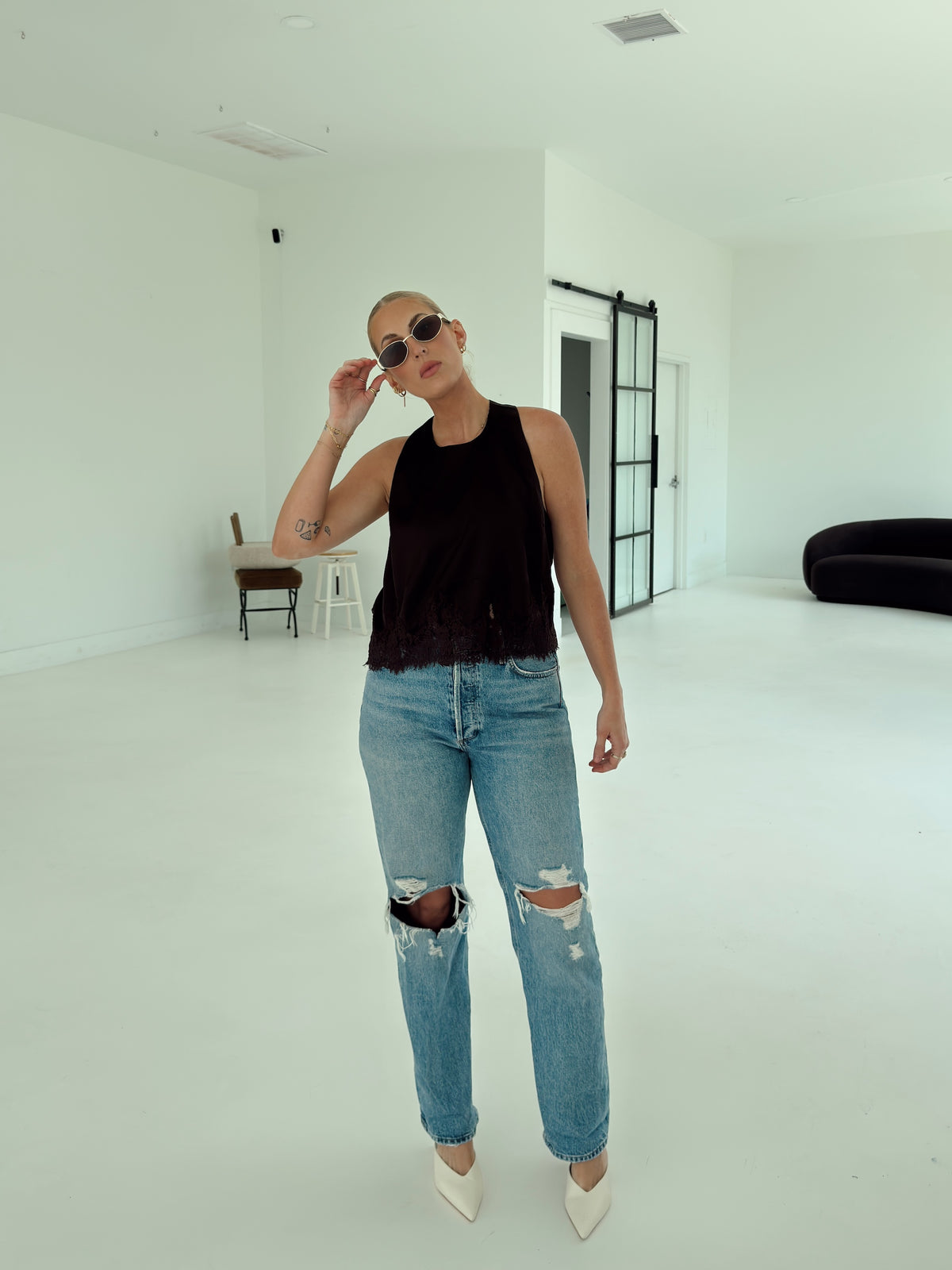 A woman stands in a bright white studio wearing a brown satin sleeveless top, light-wash distressed blue jeans with ripped knees, white pointed heels, and sunglasses.