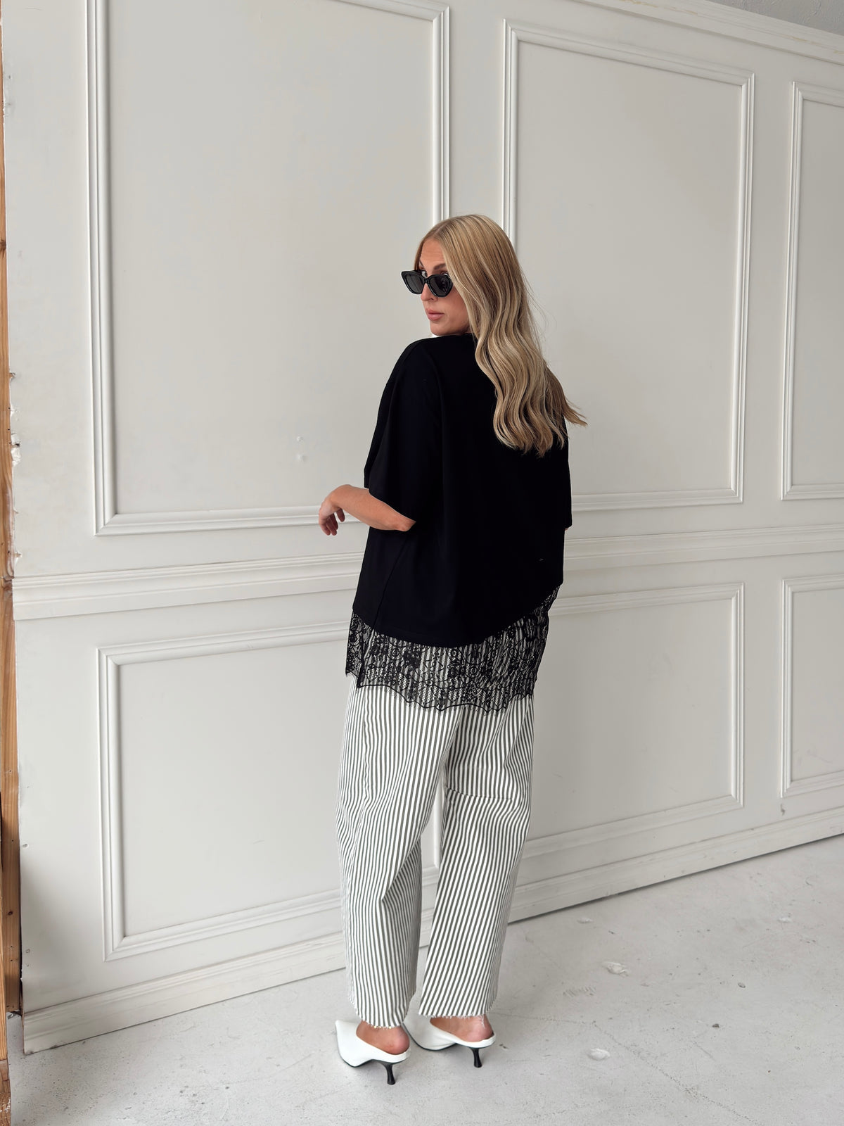 A woman modeling a pair of wide-leg barrel jeans with thin olive and white vertical stripes, paired with a black t-shirt, black sunglasses, and white pointed-toe heels, standing in front of a white paneled wall.
