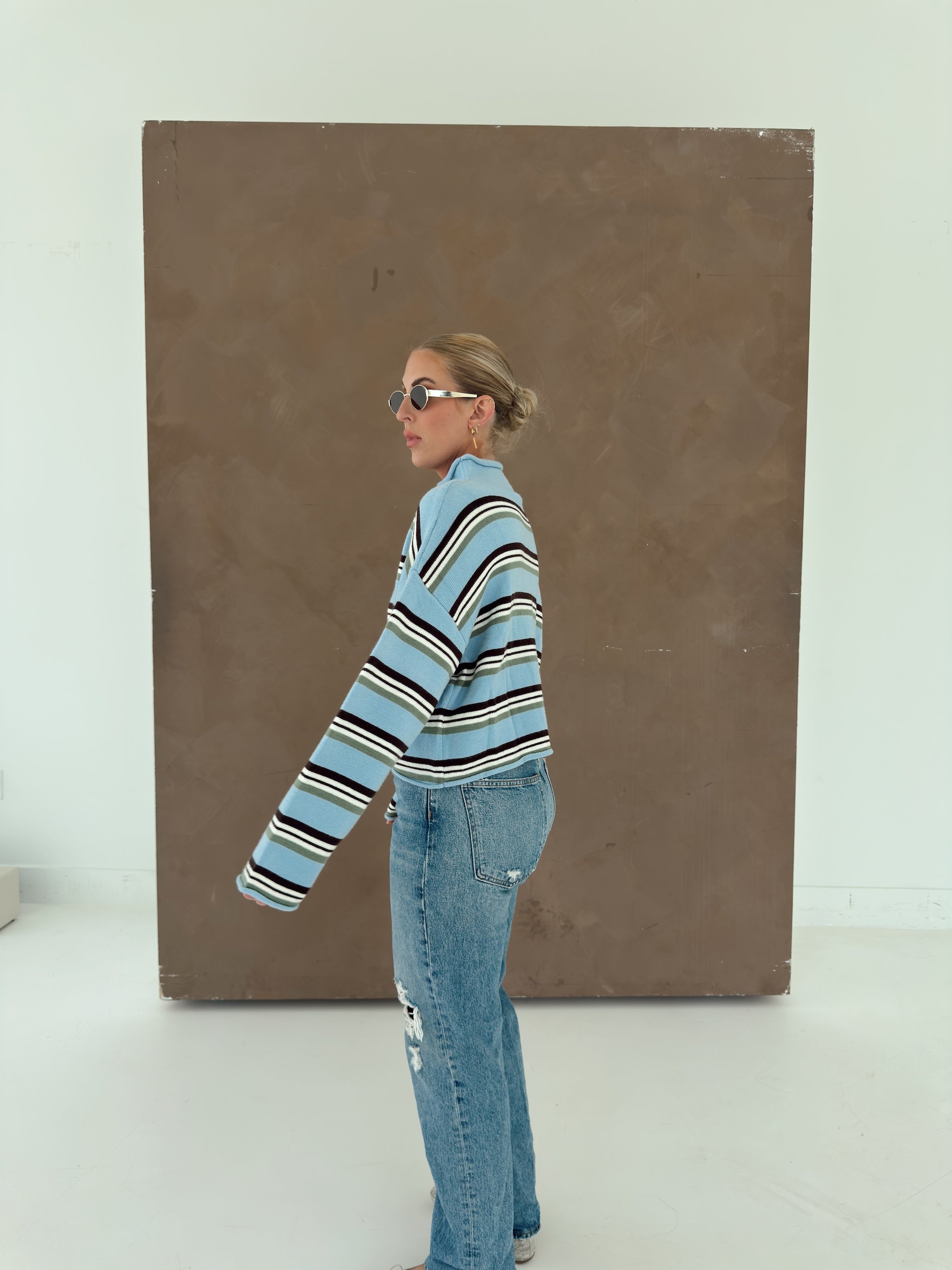 A woman wearing a light blue and brown striped knit cardigan, light-wash jeans, and sunglasses stands in profile in front of a large brown textured panel on a white studio background.