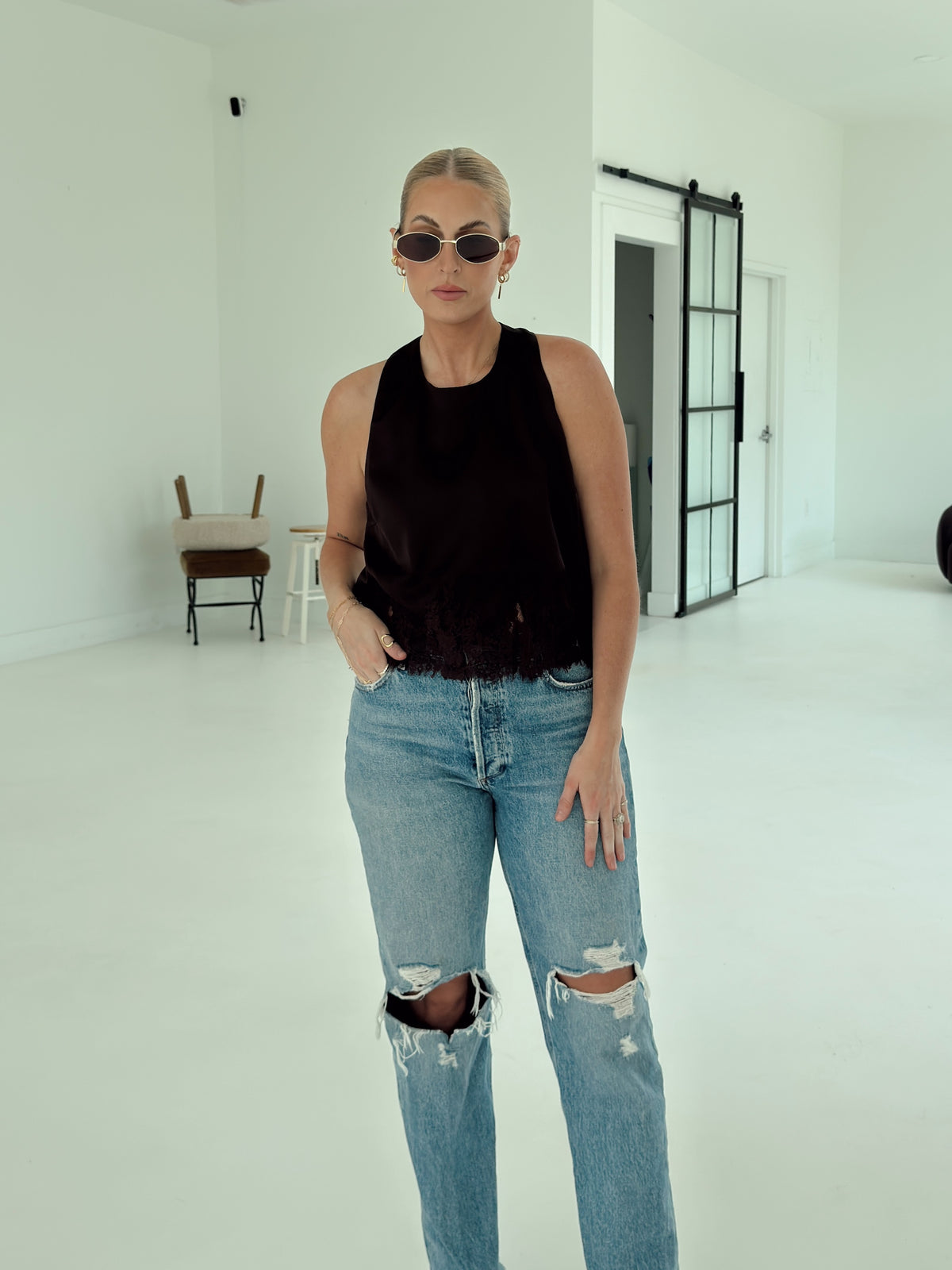 A woman modeling a sleeveless brown top and distressed light-wash blue jeans in a bright, minimalist white studio space.