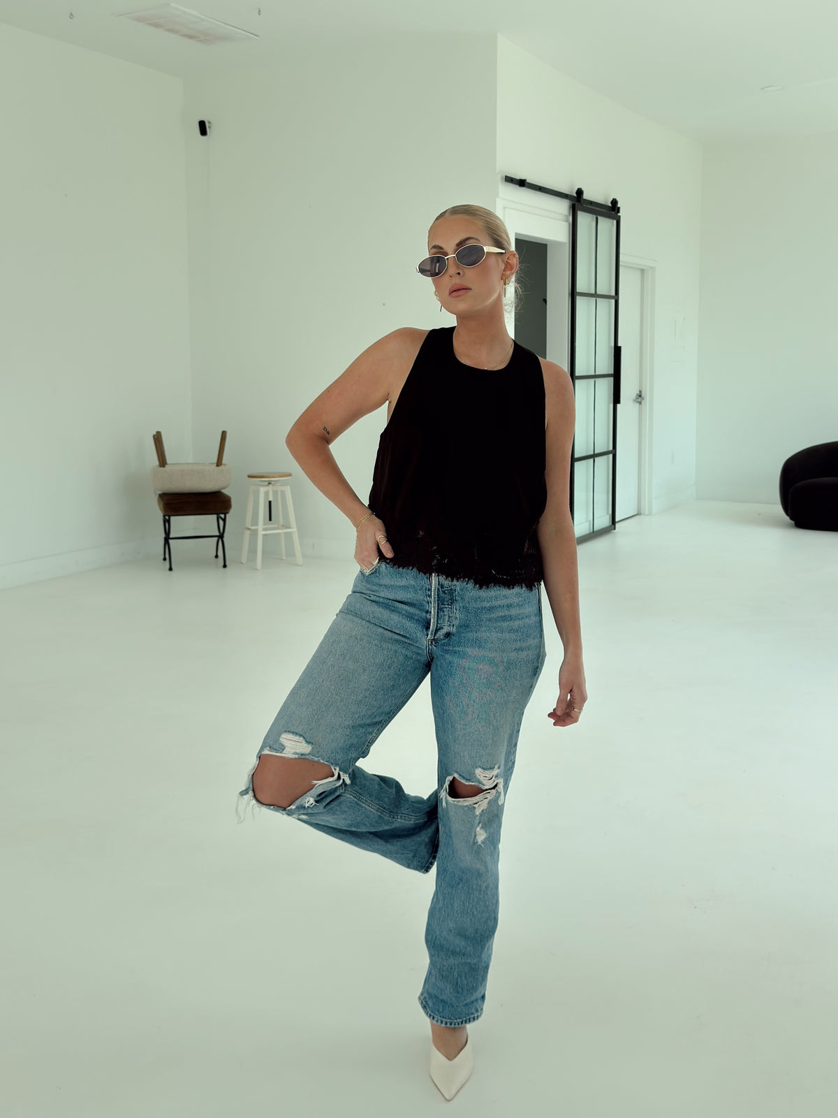 A woman posing in a minimalist white studio setting, wearing a brown sleeveless top, light-wash distressed blue jeans, and white pointed-toe heels.