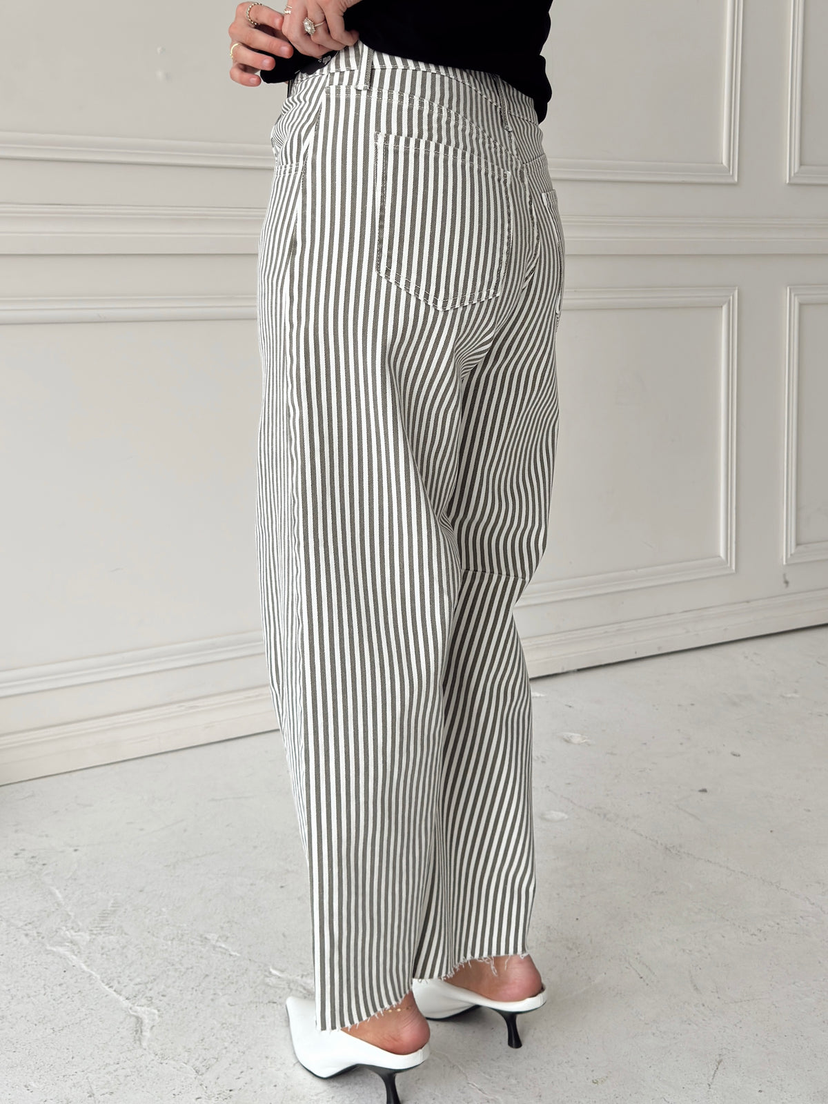 A woman modeling a pair of wide-leg barrel jeans with thin olive and white vertical stripes, paired with a black t-shirt,  and white pointed-toe heels, standing against a white paneled wall.