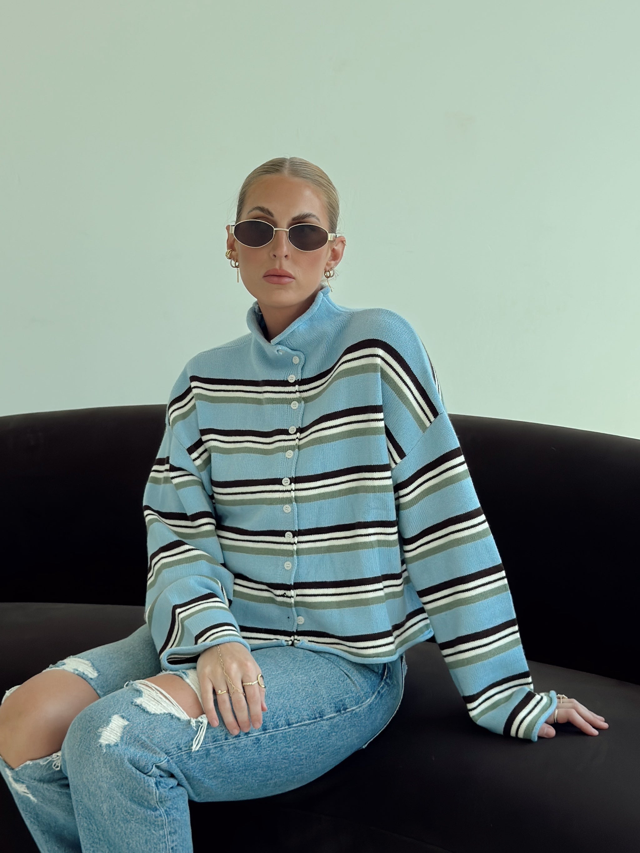 A model wearing a light blue striped button-up cardigan with brown and white stripes, paired with ripped light-wash jeans and dark sunglasses, sitting on a black couch against a pale green wall.