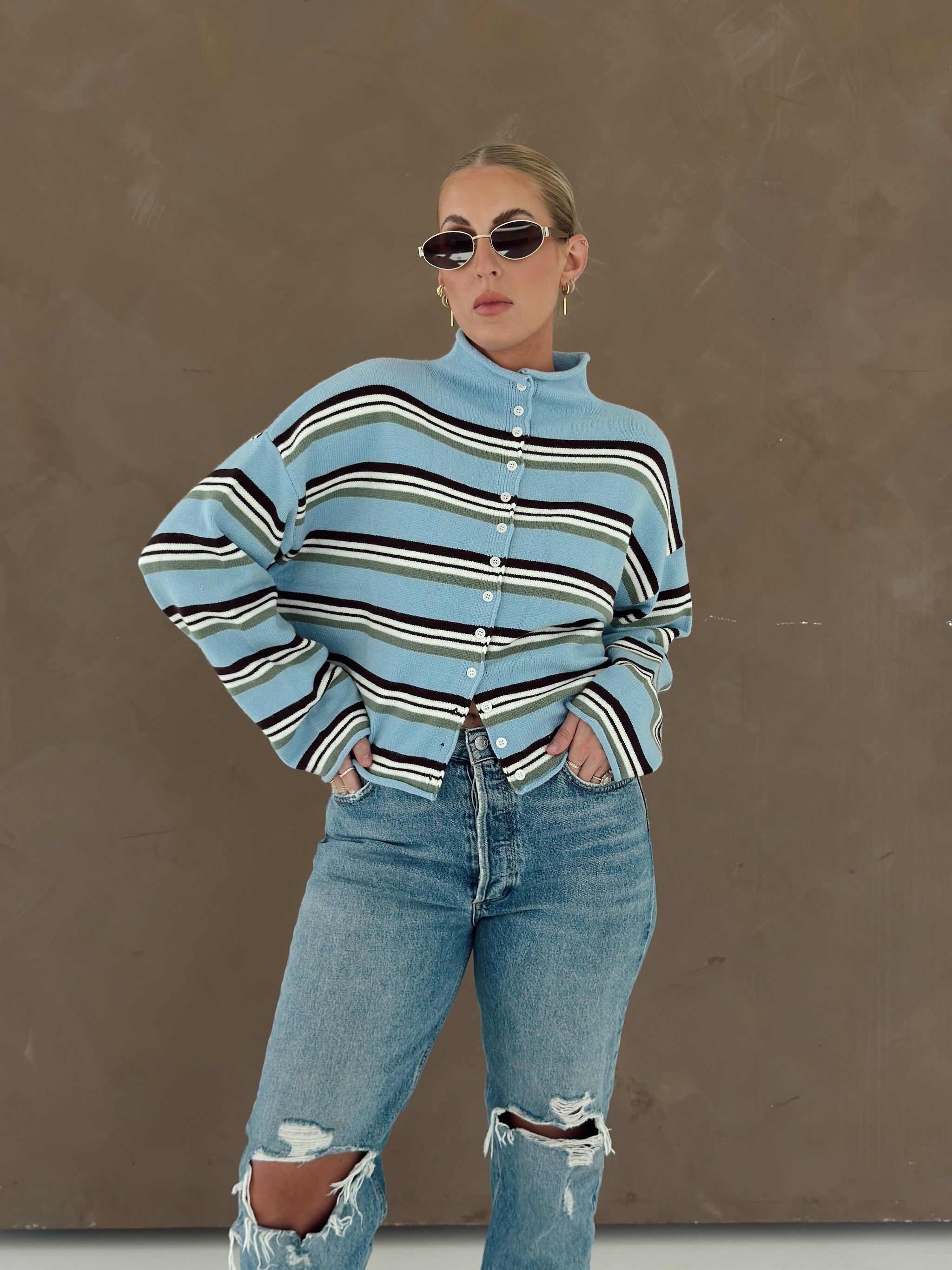 A woman modeling a light blue mock-neck button-up sweater with brown, white, and olive green horizontal stripes, paired with distressed blue jeans, against a textured brown wall.