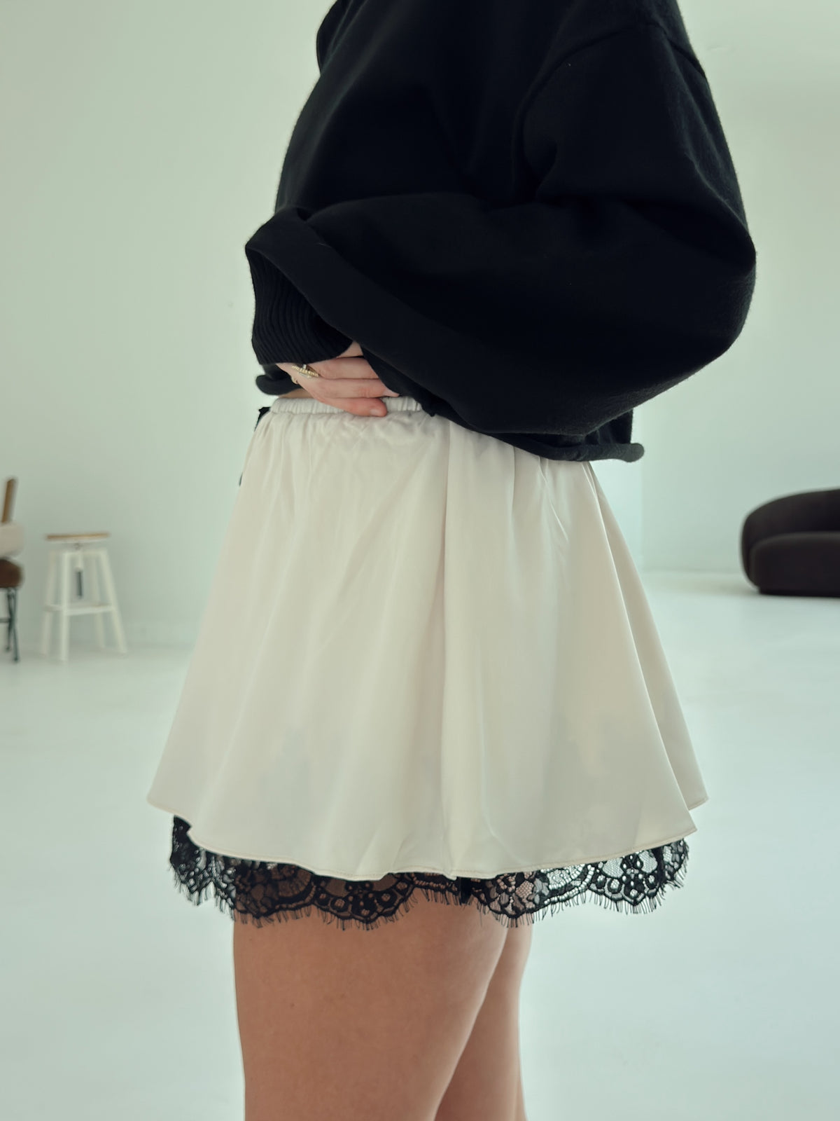 A person wearing a black oversized sweatshirt and a cream-colored mini skirt with a black lace hem, standing in a minimalist white room.