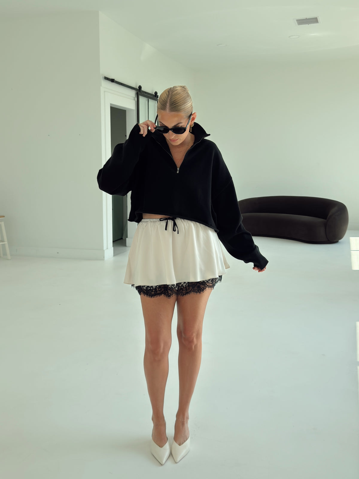 A woman stands in a bright white room wearing a black cropped zip-up hoodie, a cream mini skirt with black lace trim, and white pointed-toe heels.