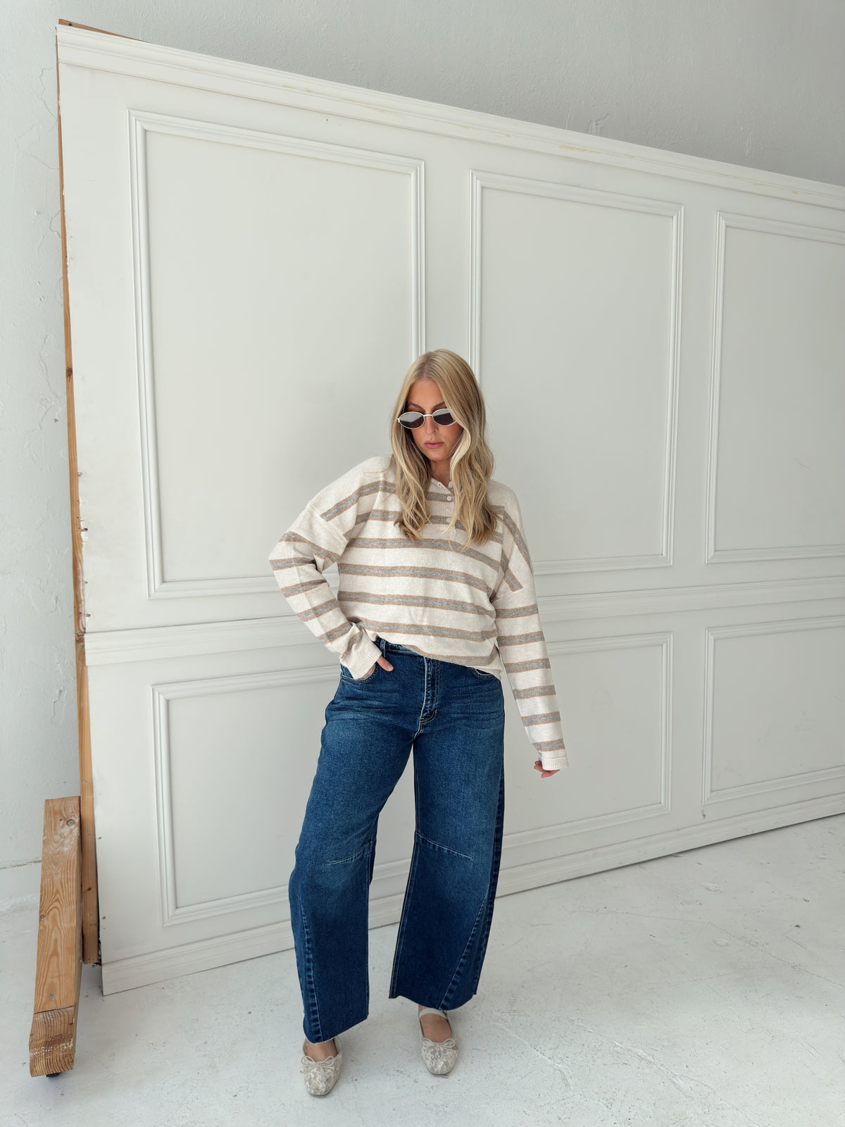 A woman stands in front of a white paneled wall wearing a beige, cream, and light blue horizontally striped long-sleeve knit top, wide-leg blue jeans, and light-colored flats.