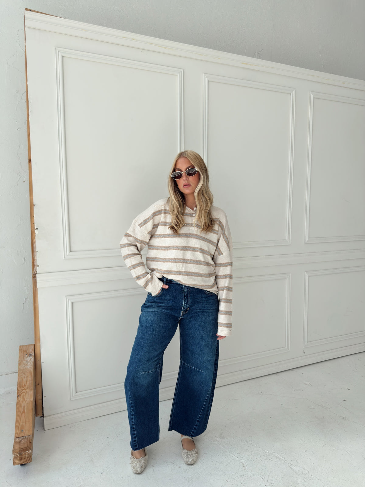 A woman stands in front of a white paneled wall wearing a beige, cream, and light blue horizontally striped long-sleeve knit top, wide-leg blue jeans, and light-colored flats.