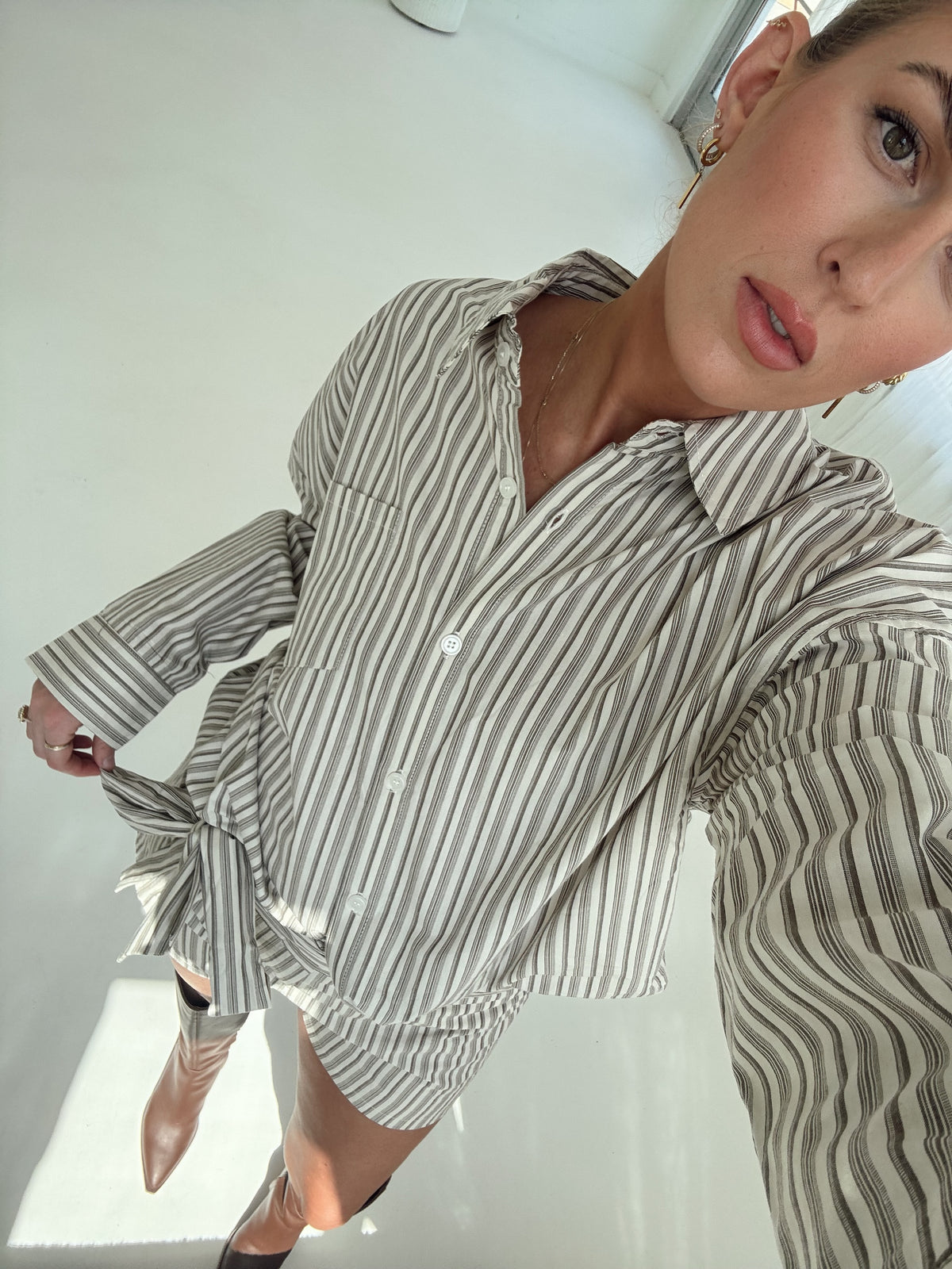 A woman wearing a brown and white vertically striped button-down shirt with a tie waist, photographed in a bright, minimalist indoor setting.