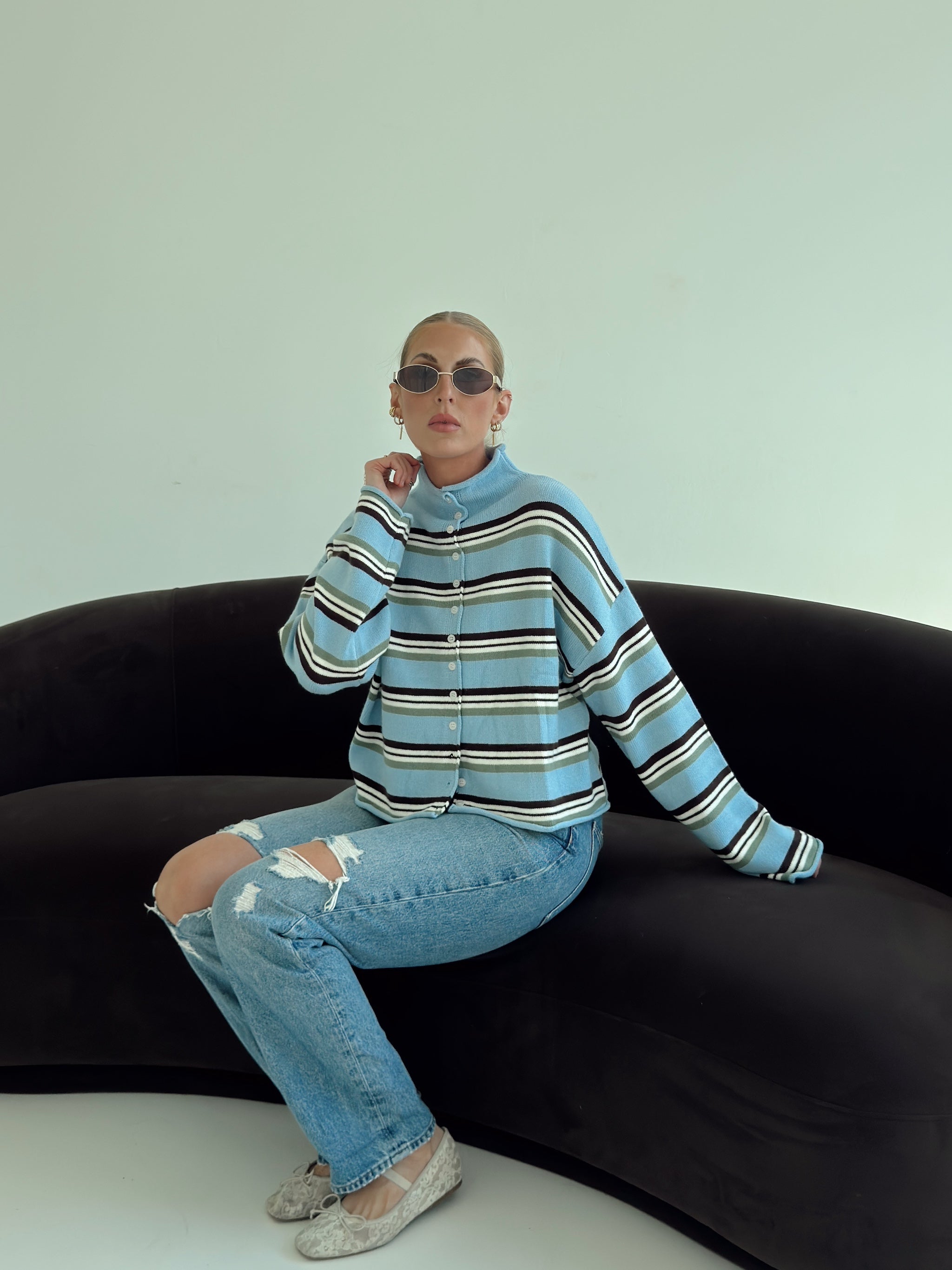 A model wearing a light blue and brown striped knit turtleneck sweater, distressed light-wash jeans, and sunglasses, sitting on a black curved sofa against a pale mint green wall.