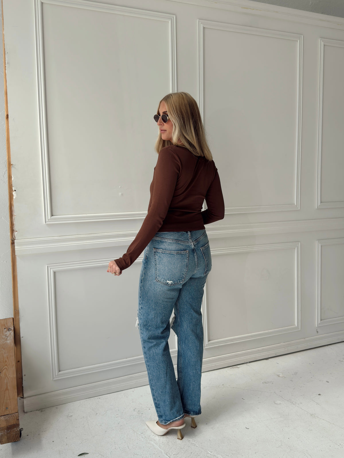 A woman modeling a brown long sleeve fitted top with light blue jeans, sunglasses, and white mule heels.