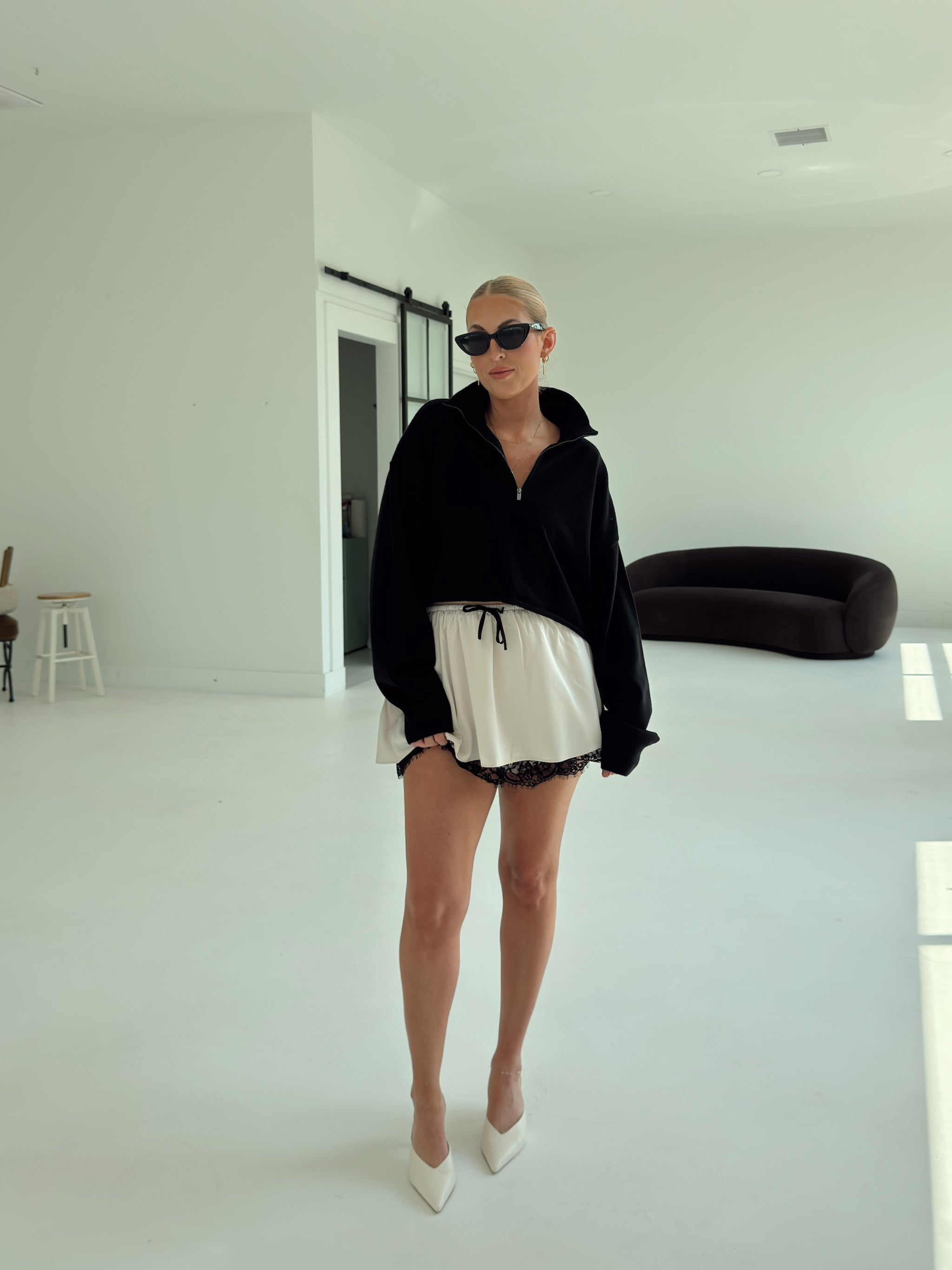 A woman stands in a bright, minimalist white room wearing a black half-zip sweatshirt, a cream ruffled mini skirt with black lace trim, and white pointed-toe heels.