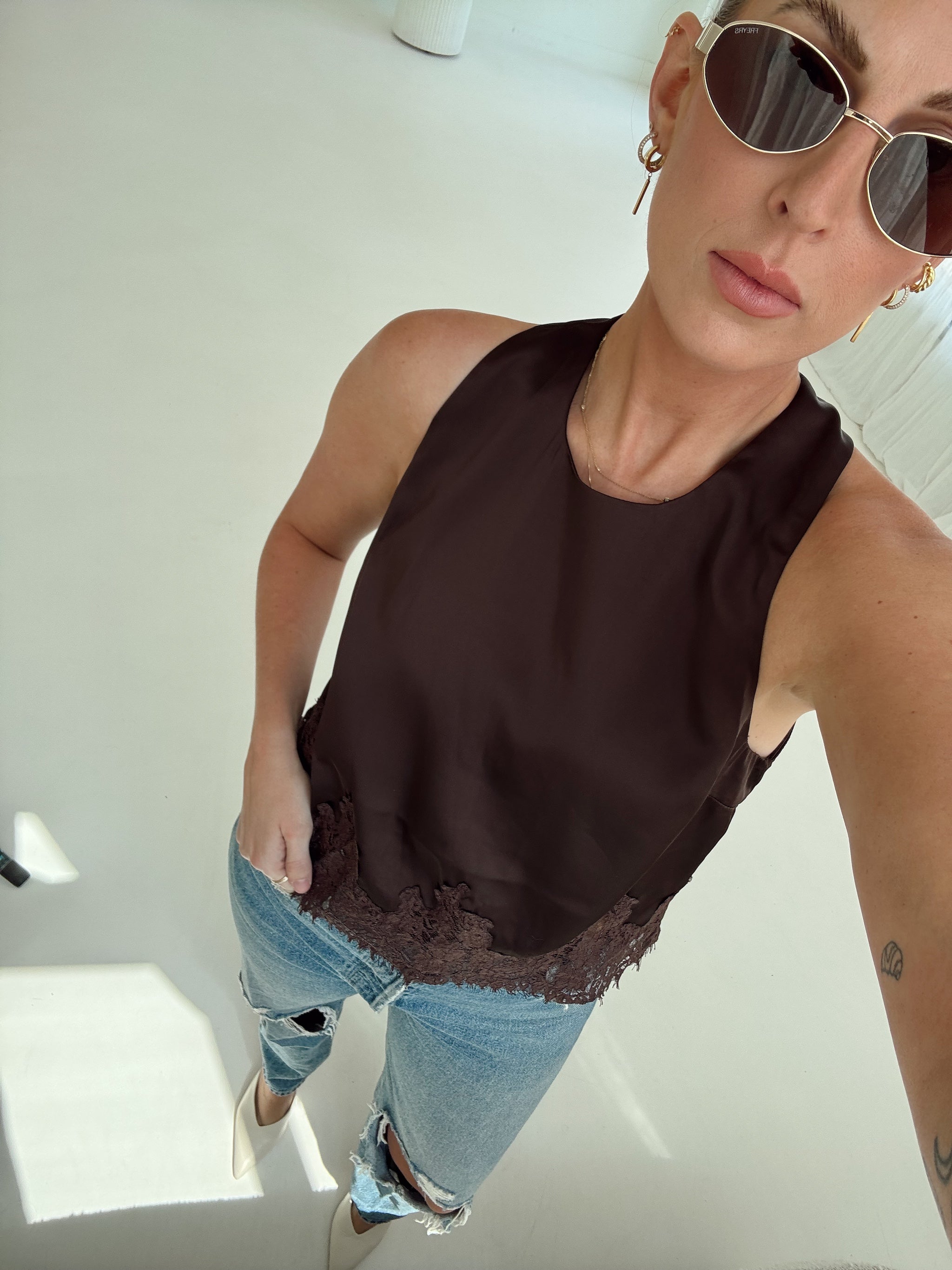 A woman wearing a sleeveless dark brown top with a lace hem, paired with light-wash distressed jeans and sunglasses, captured in a bright, minimalist indoor setting.