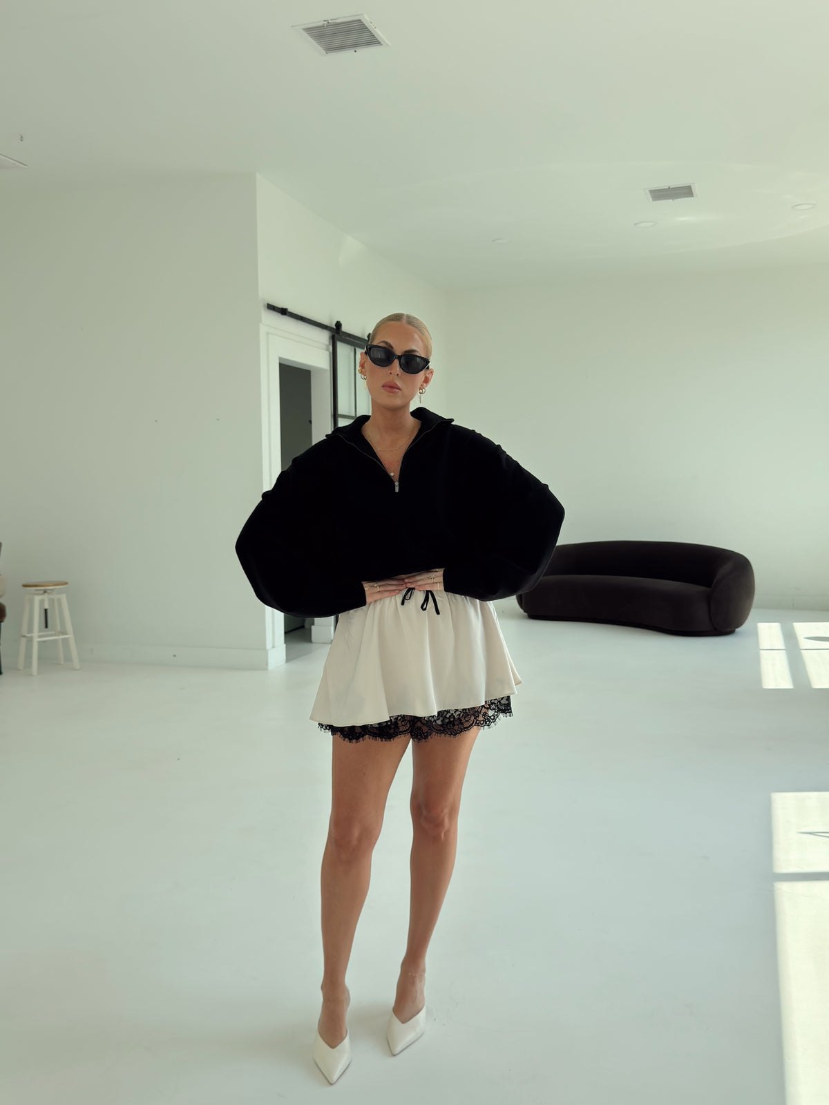 A woman stands in a bright, minimalist white room wearing a black oversized zip-up hoodie with voluminous sleeves, a cream mini skirt with black lace trim, and white pointed-toe heels.