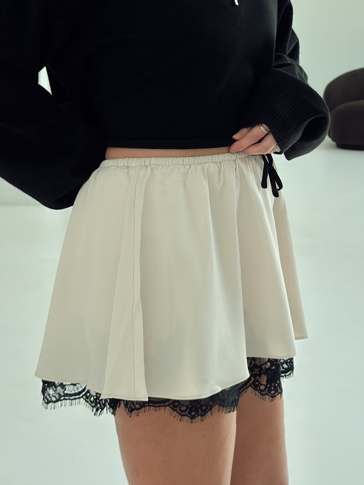 An off-white or cream flared mini skirt with black lace trim at the hem and a black drawstring tie at the elastic waistband, shown on a model wearing a black long-sleeve top.