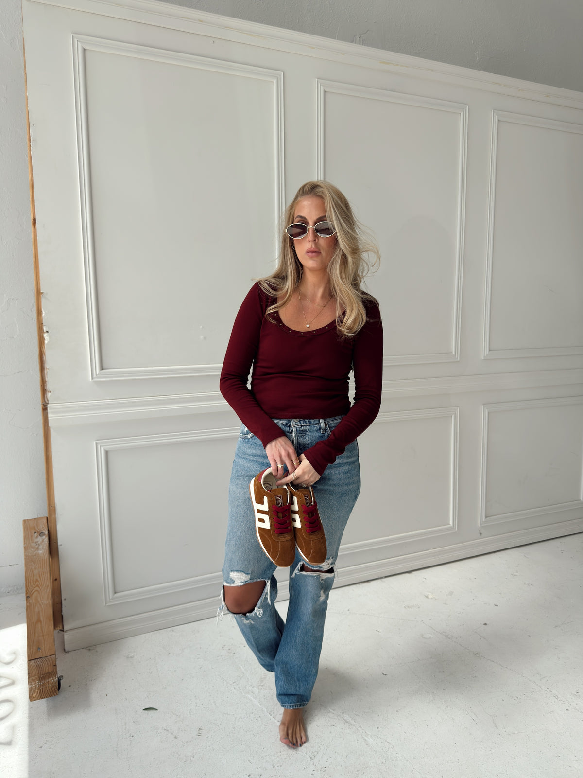 A woman stands barefoot in a white paneled room, wearing a burgundy long-sleeve top, ripped blue jeans, and sunglasses while holding a pair of brown shoes with white and red details.