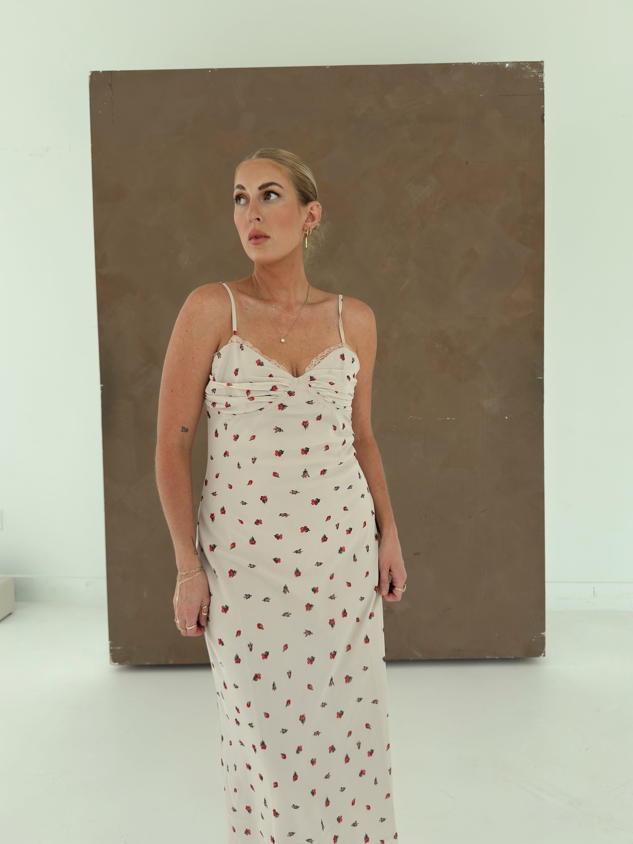 A woman modeling a beige spaghetti-strap maxi dress with a small red floral print, standing in front of a large brown canvas painting in a white studio.