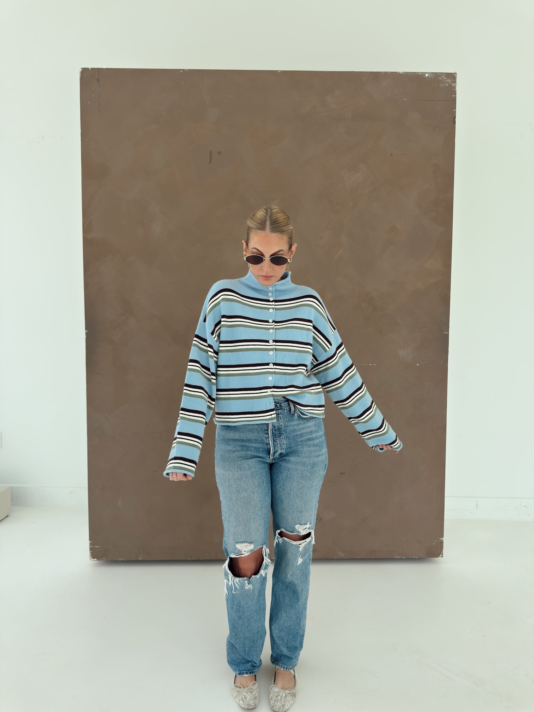 A person wearing a light blue knit cardigan with horizontal brown, white, and olive green stripes, paired with light-wash jeans and lace flats.