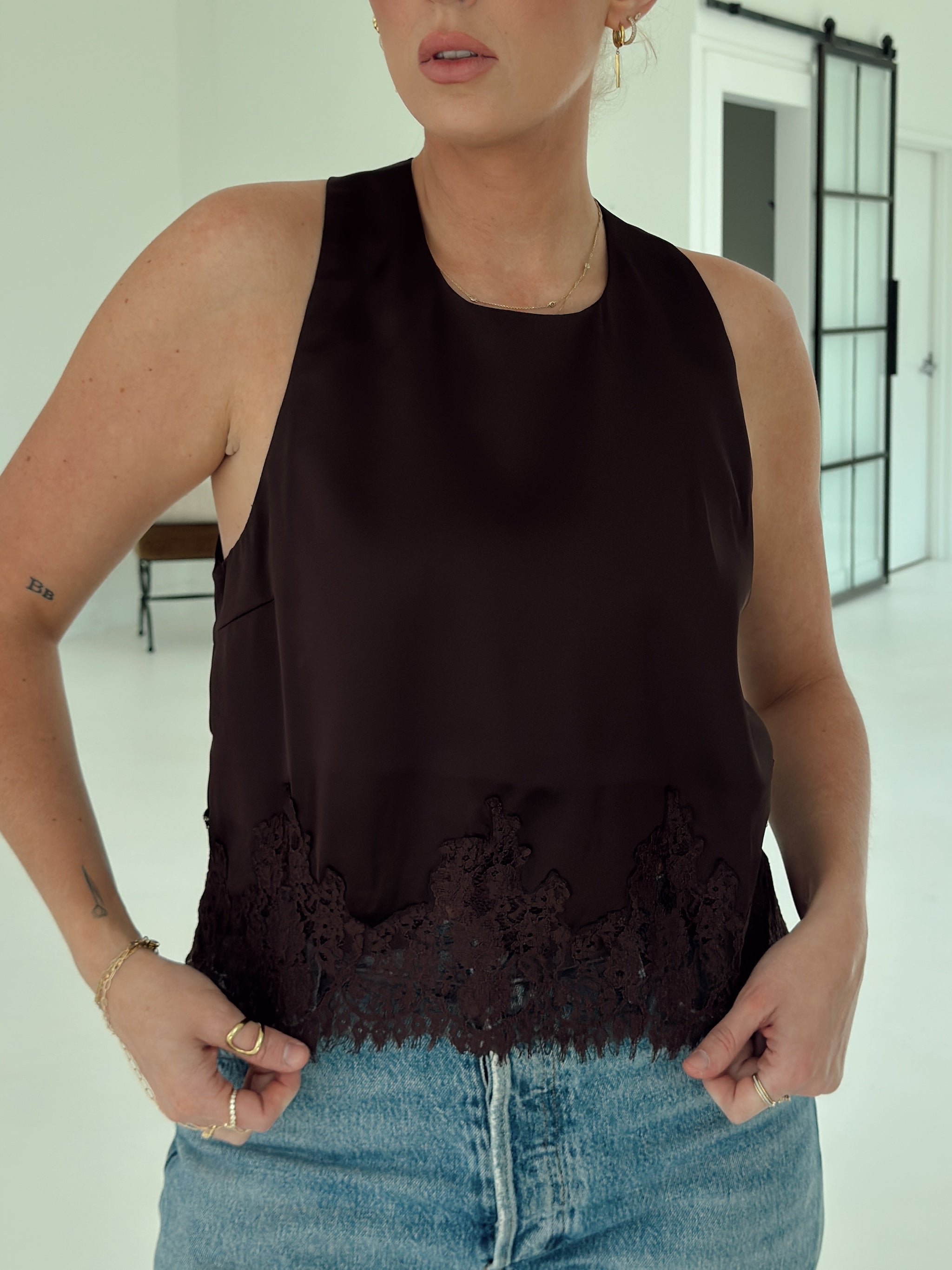 A woman wearing a sleeveless dark brown top with a scalloped lace hem, paired with light blue denim jeans, in a bright indoor room.