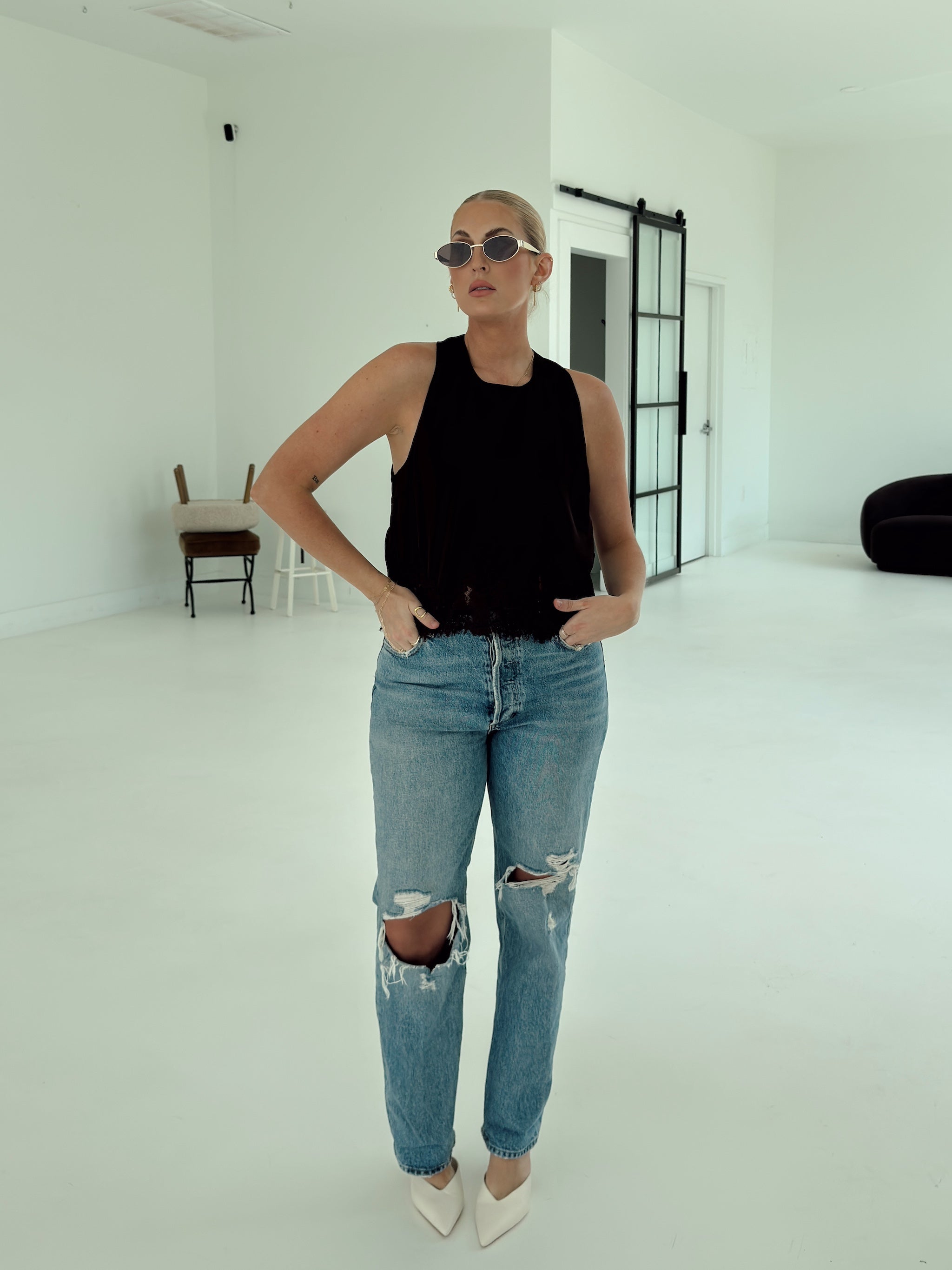 A woman posing in a minimalist white studio setting, wearing a brown sleeveless top, light-wash distressed blue jeans, and white pointed-toe heels.