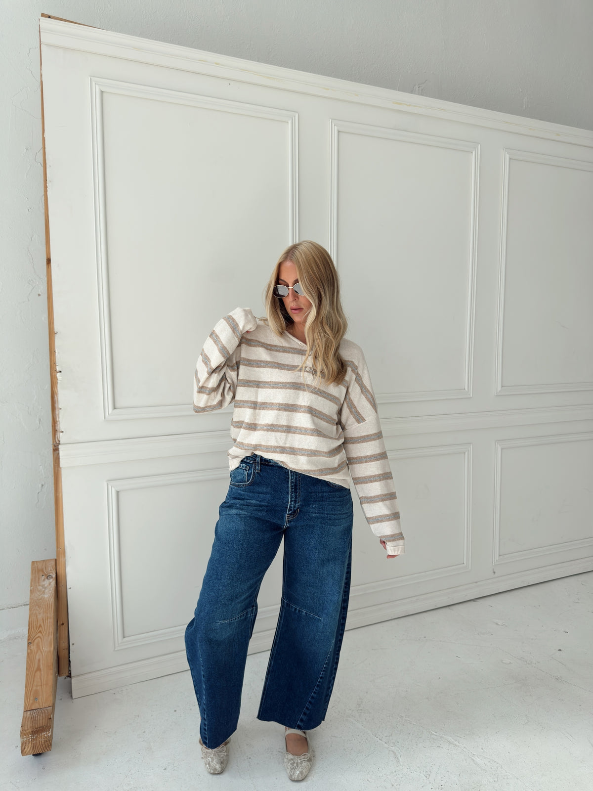 A woman stands in front of a white paneled wall wearing a beige, cream, and light blue horizontally striped long-sleeve knit top, wide-leg blue jeans, and light-colored flats.