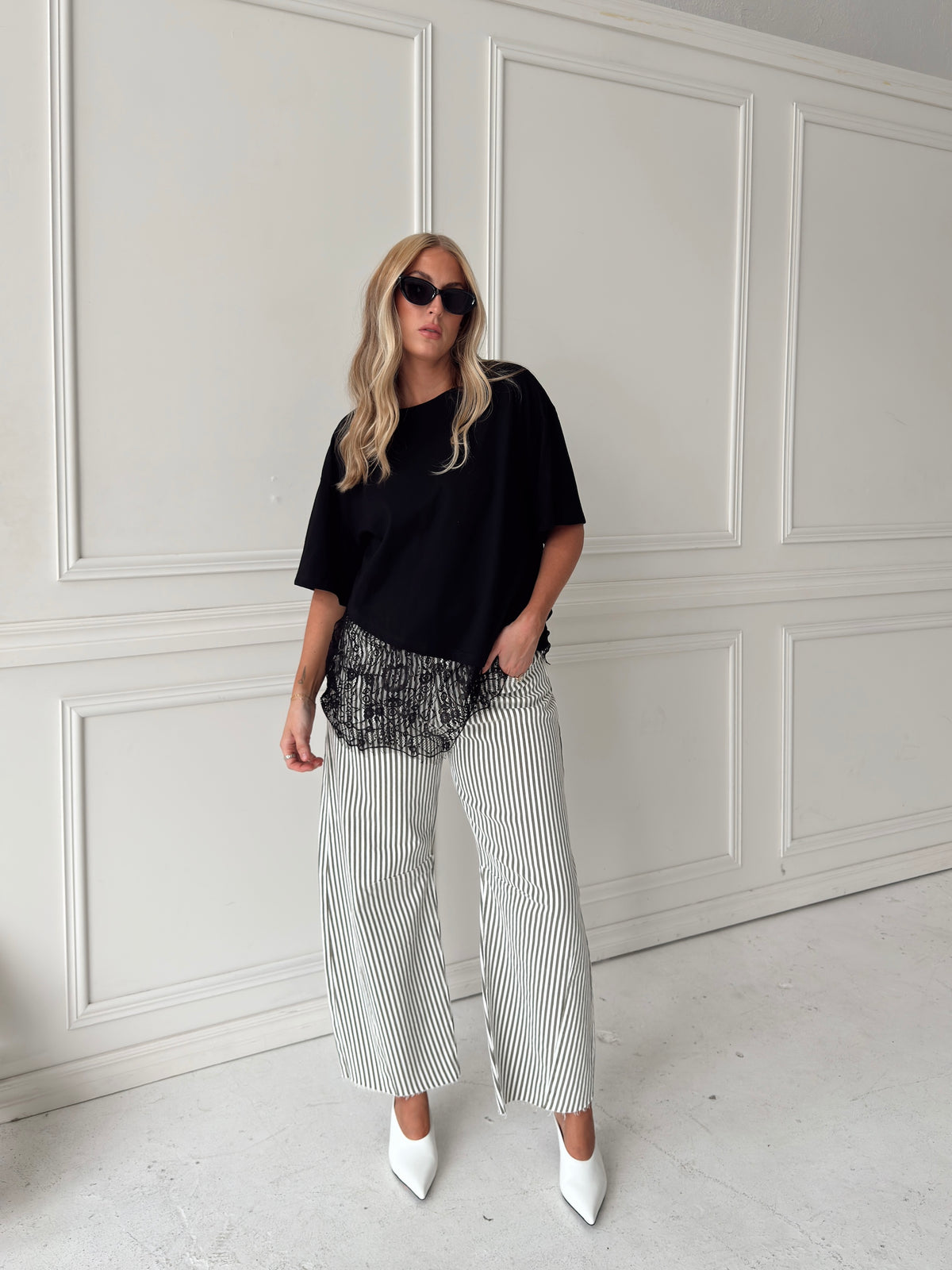 A woman modeling a pair of wide-leg barrel jeans with thin olive and white vertical stripes, paired with a black t-shirt, black sunglasses, and white pointed-toe heels, standing in front of a white paneled wall.