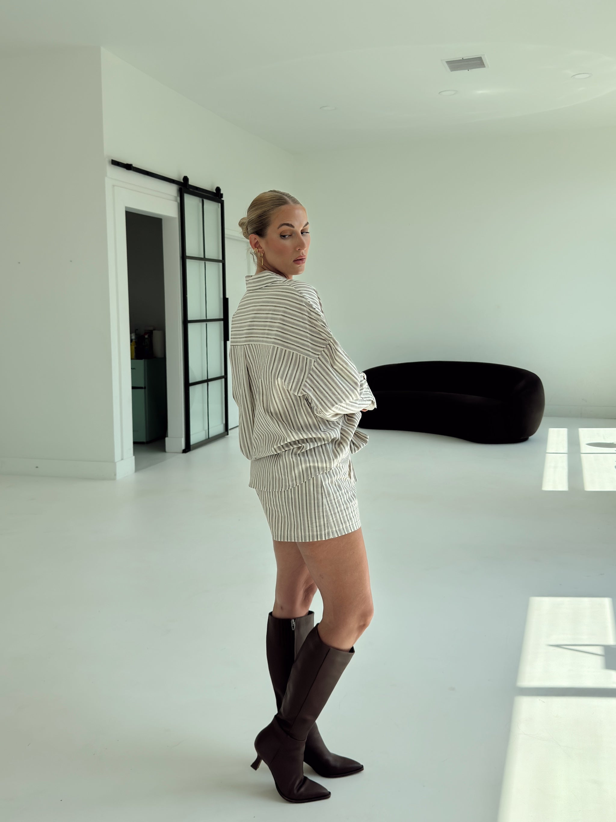 A woman stands in a bright, minimalist white room wearing a cream and white vertically striped short-sleeve shirt and matching striped shorts, paired with dark brown pointed-toe heeled boots.