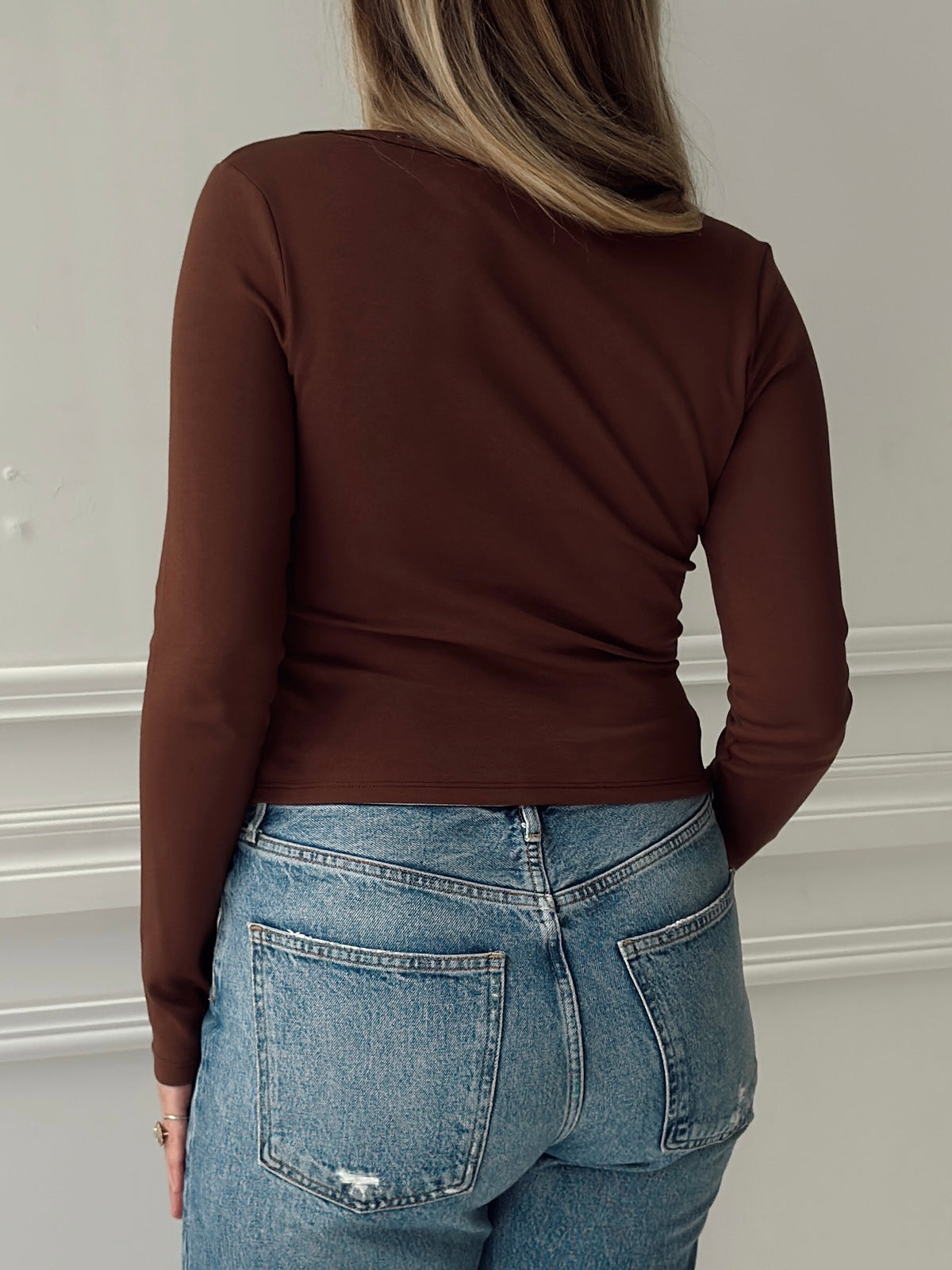 A woman shown from the back wearing a fitted long-sleeve brown top and light blue jeans against a white wall with white molding.