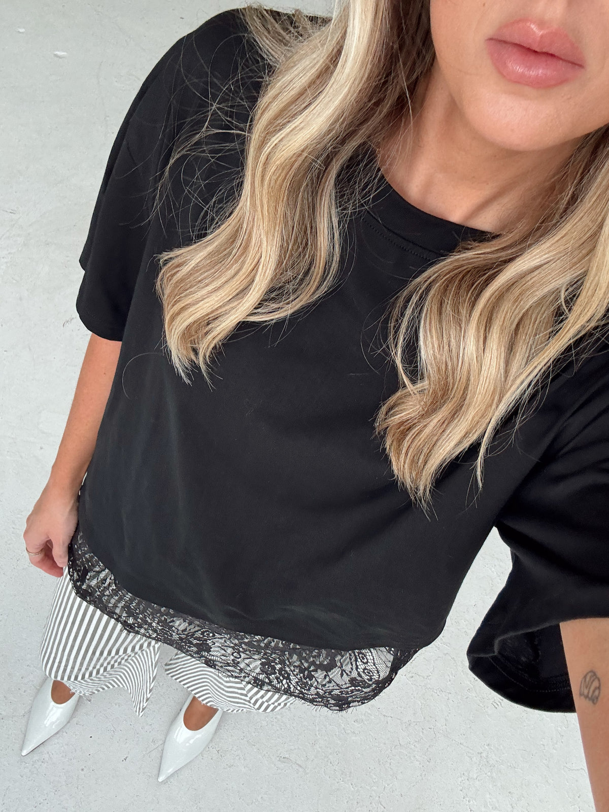 A woman wearing a black oversized short-sleeve t-shirt with a sheer black lace hem, styled with olive and white striped pants and white pointed-toe heels.