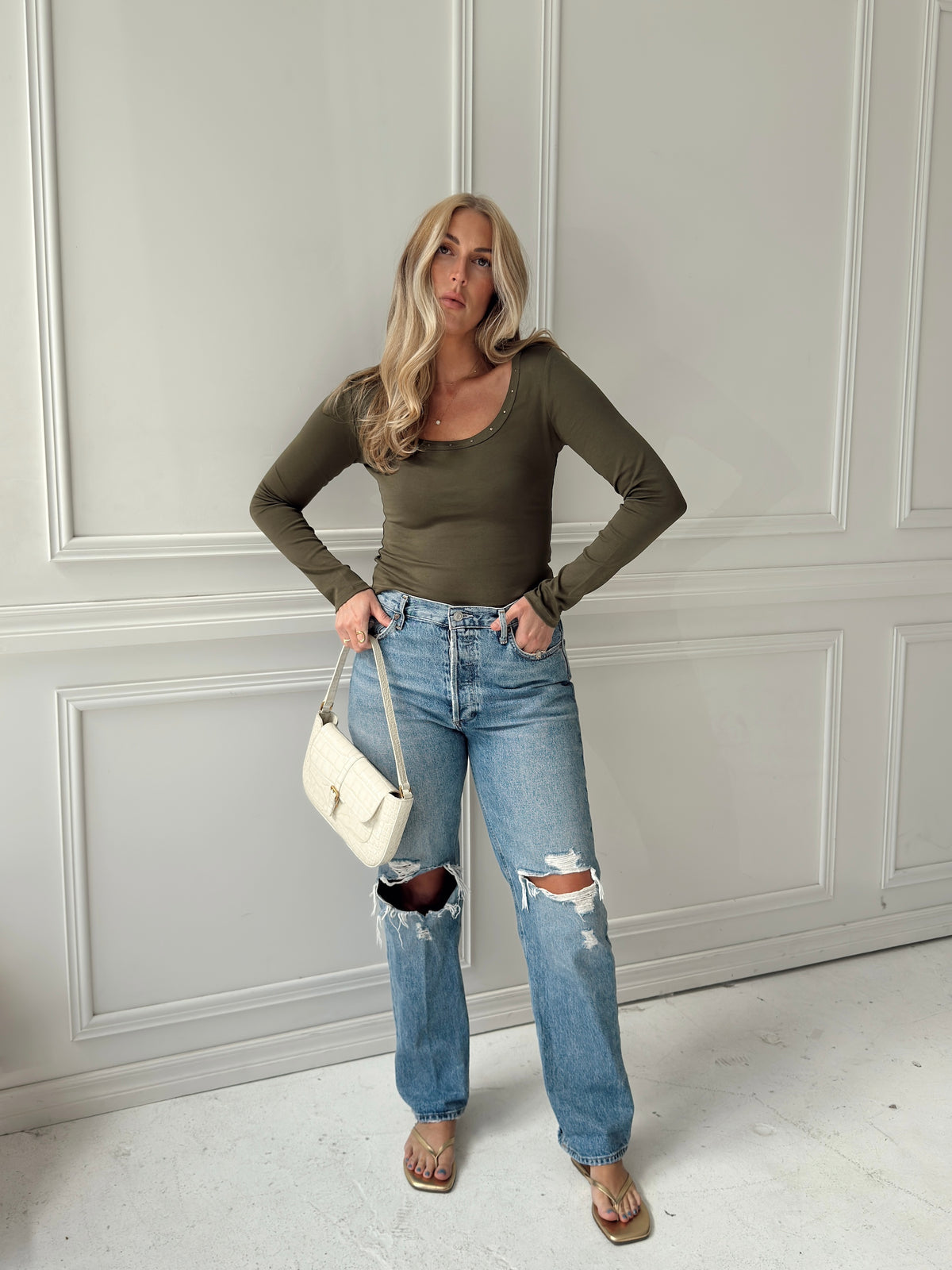 A woman stands in a room with white paneled walls wearing an olive green long-sleeve scoop-neck top, distressed light-wash blue jeans, and sandals, while holding a small white handbag.