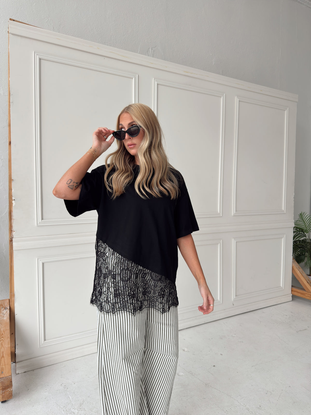 A woman modeling a black short-sleeve top with a sheer black lace overlay and wide-leg olive and white striped pants, standing in front of a white paneled wall.