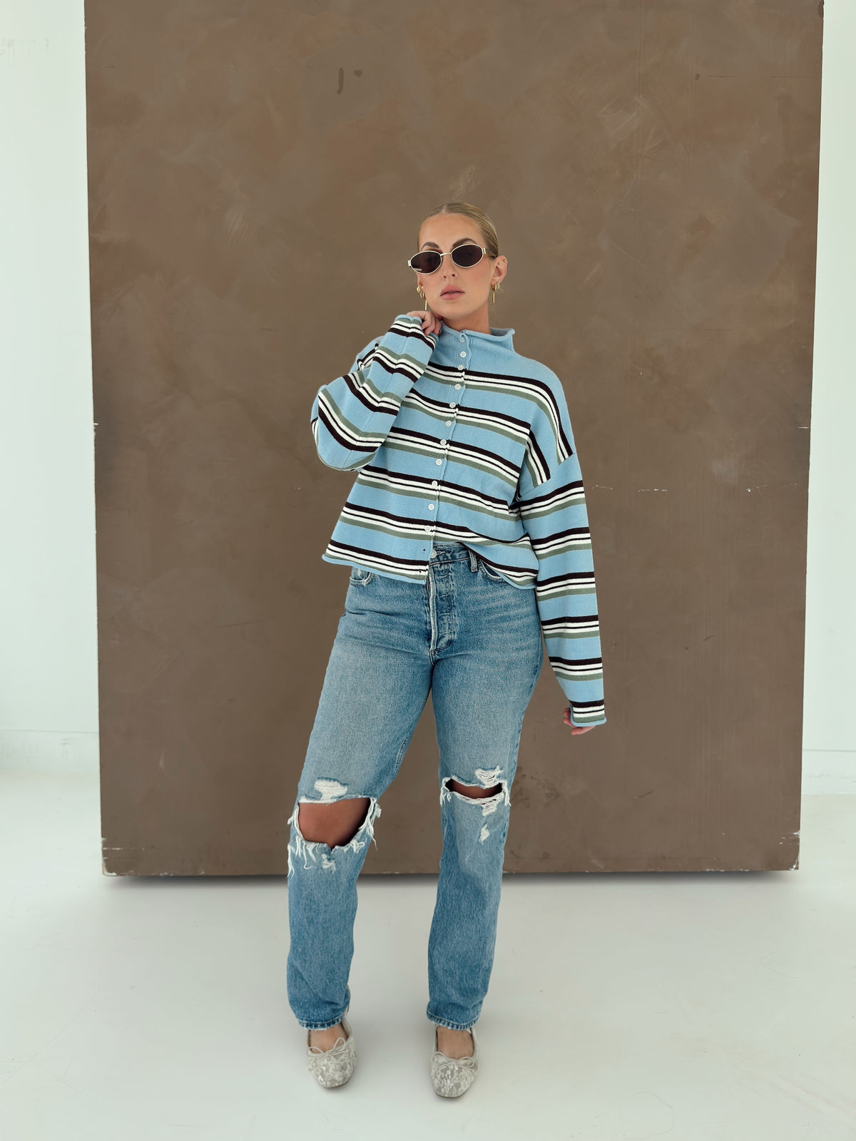 A woman modeling a light blue  cardigan with brown, ivory and green stripes with distressed blue jeans and lace shoes, standing in front of a large brown textured backdrop.