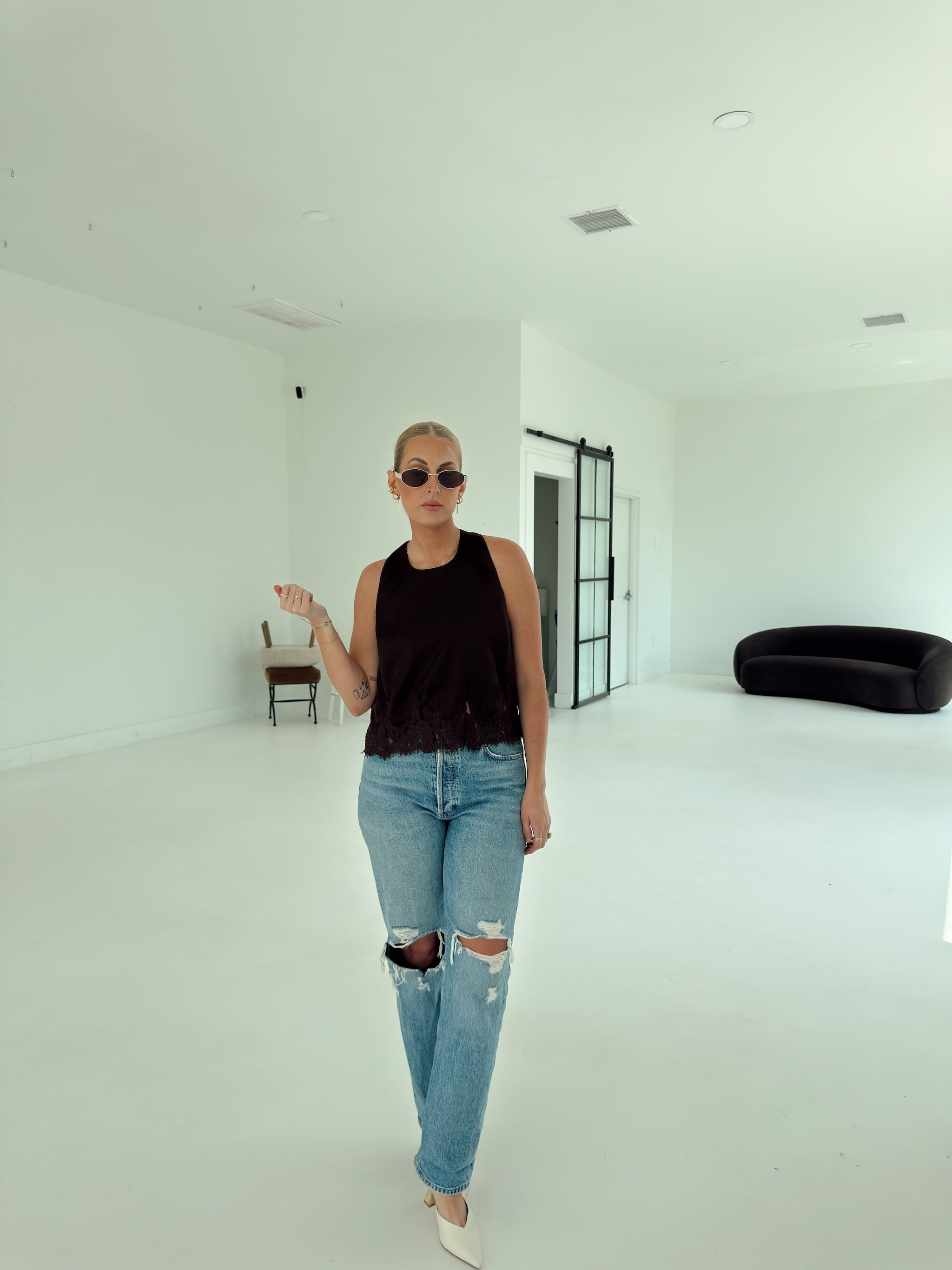 A woman wearing a brown sleeveless ruffled top, distressed light-wash blue jeans, and white pointed shoes walks through a bright, minimalist white studio space.