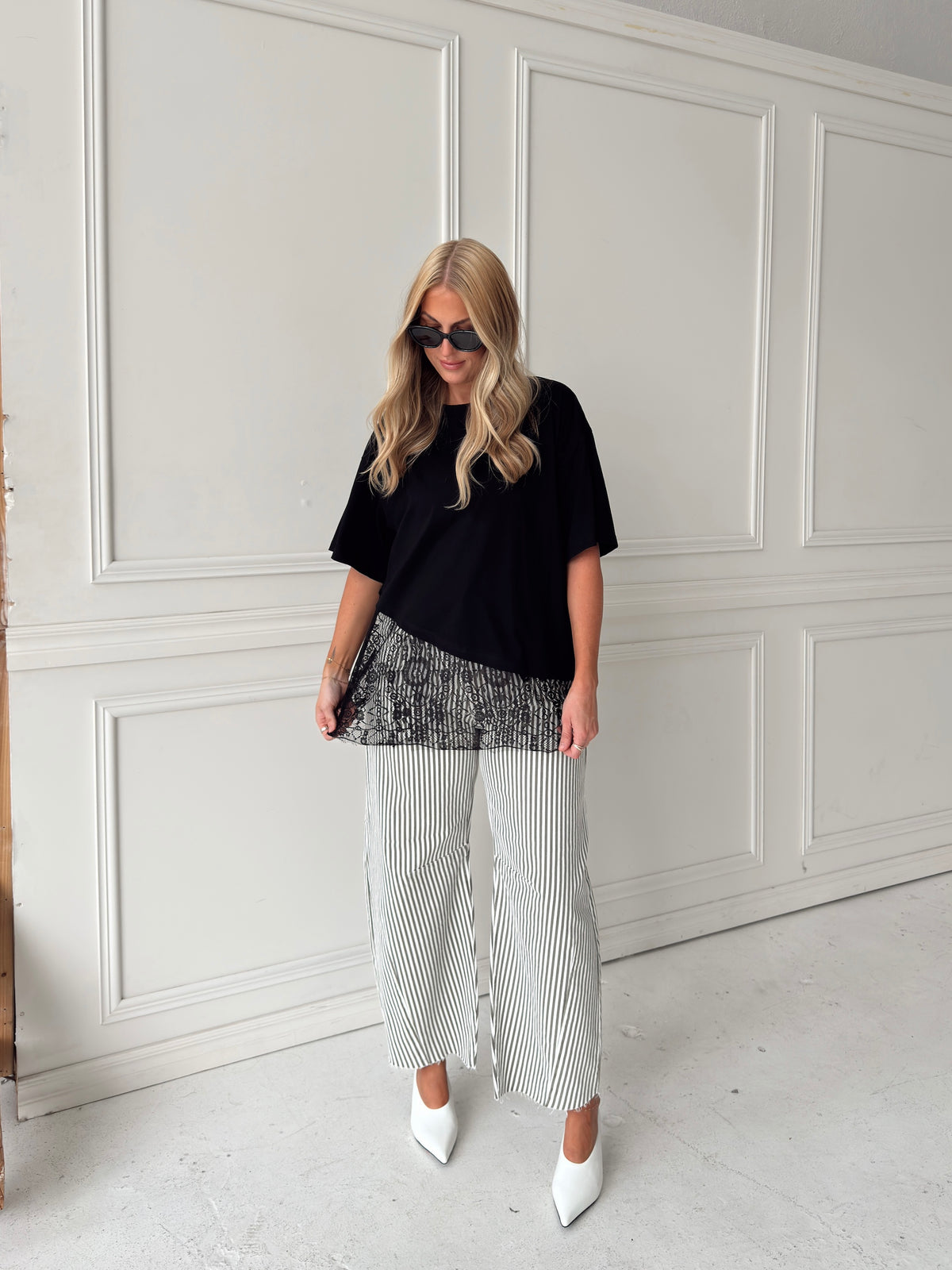 A woman modeling a black asymmetrical top with a sheer black lace hem, paired with white and olive vertically striped pants, standing in front of a white paneled studio backdrop.