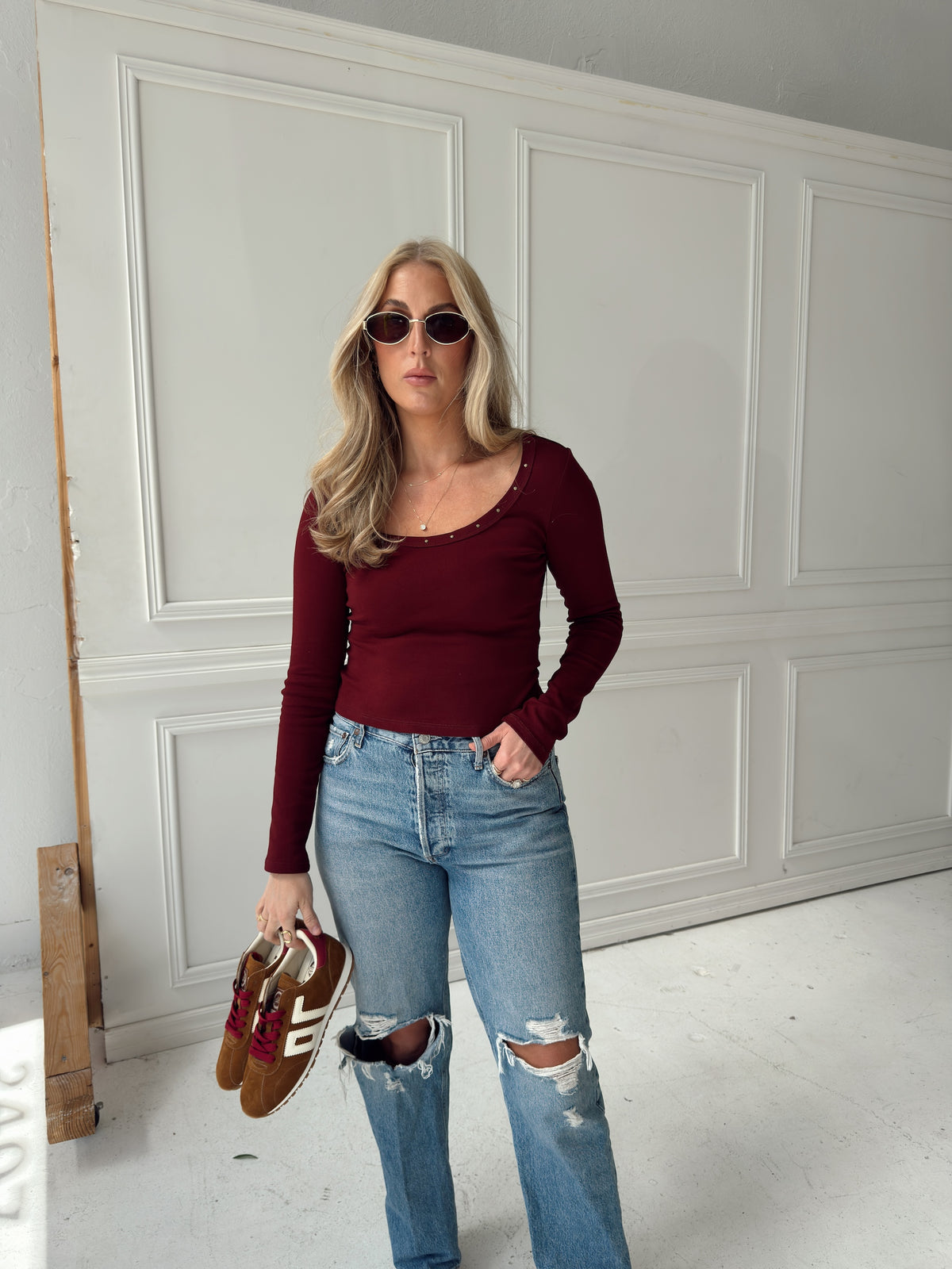 A woman wearing a burgundy long-sleeve scoop-neck top, light-wash distressed jeans, and sunglasses, holding a pair of brown sneakers in front of a white paneled wall.