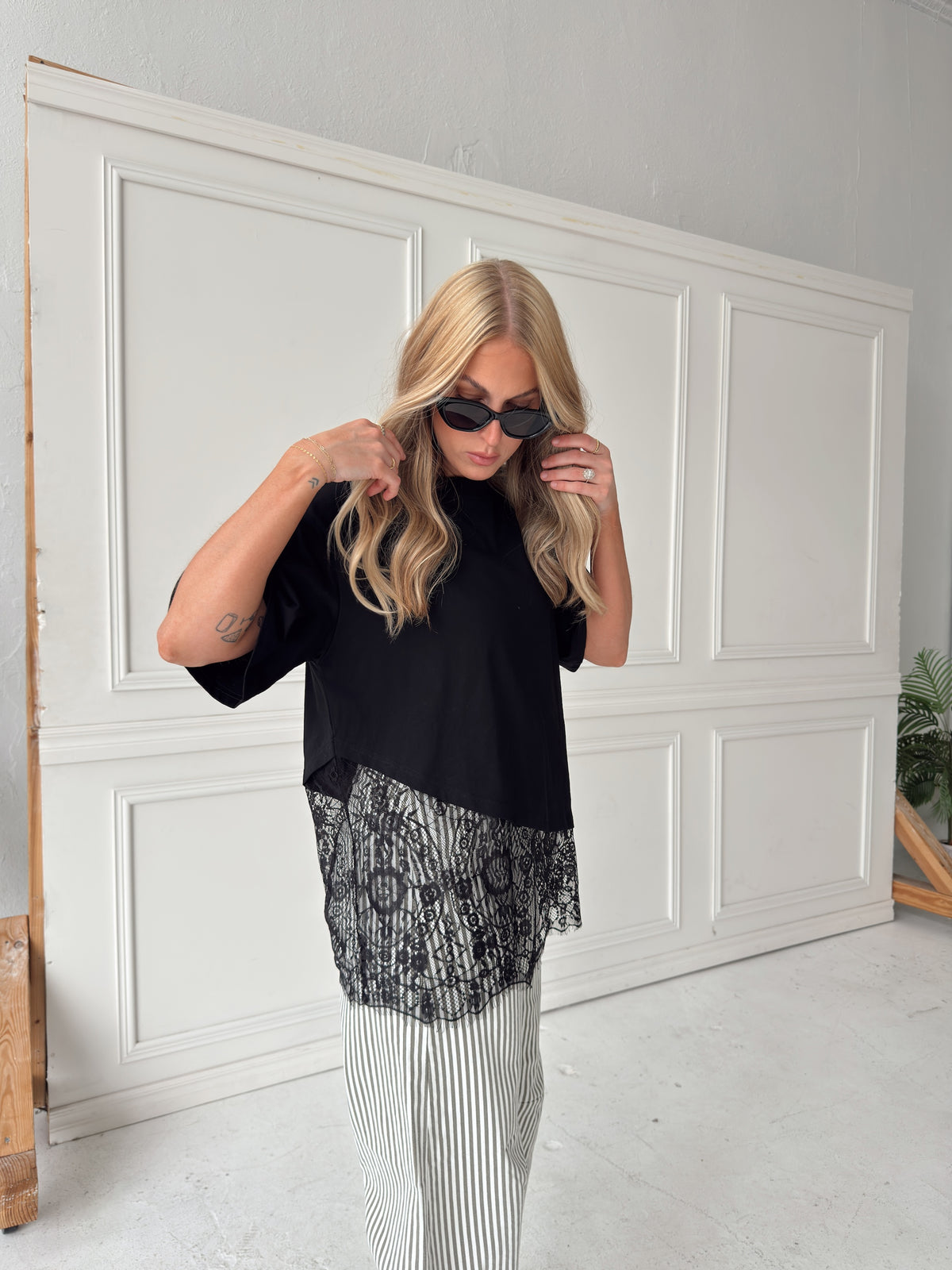 A woman modeling a black short-sleeve top with a sheer black lace hem overlay with olive and white vertically striped pants, standing in front of a white paneled wall.